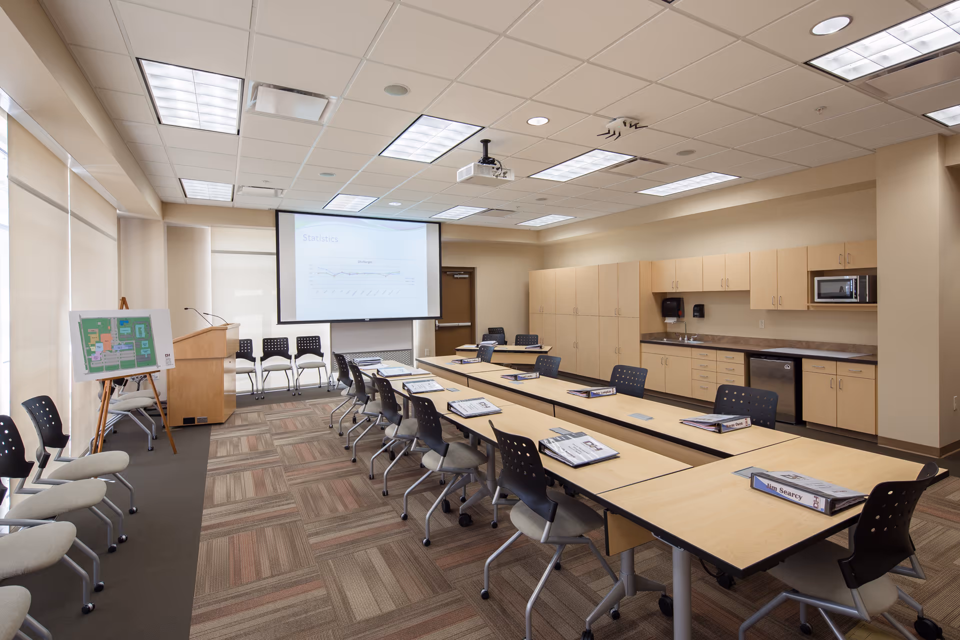 A modern training room in a healthcare facility with long desks, chairs, projector, and kitchenette.