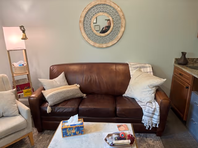Cozy living room with brown leather sofa, pillows, round wall mirror, and kitchenette.