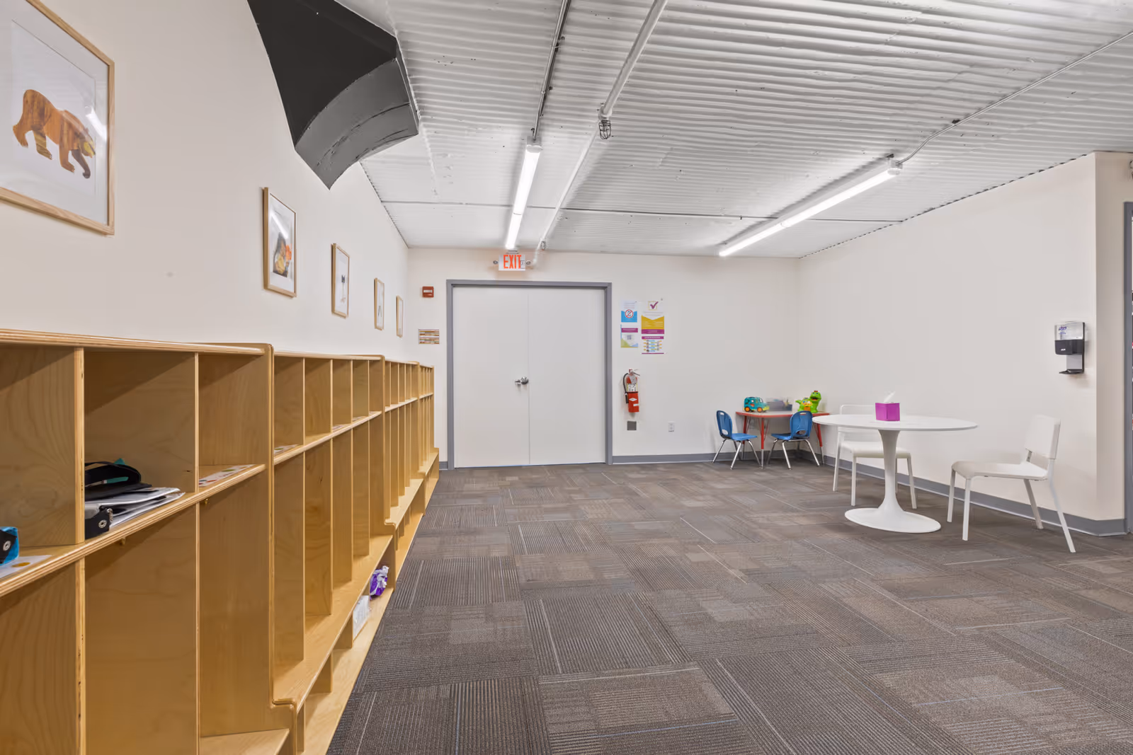 An empty activity room with cubby shelves, framed animal pictures, and a round white table.