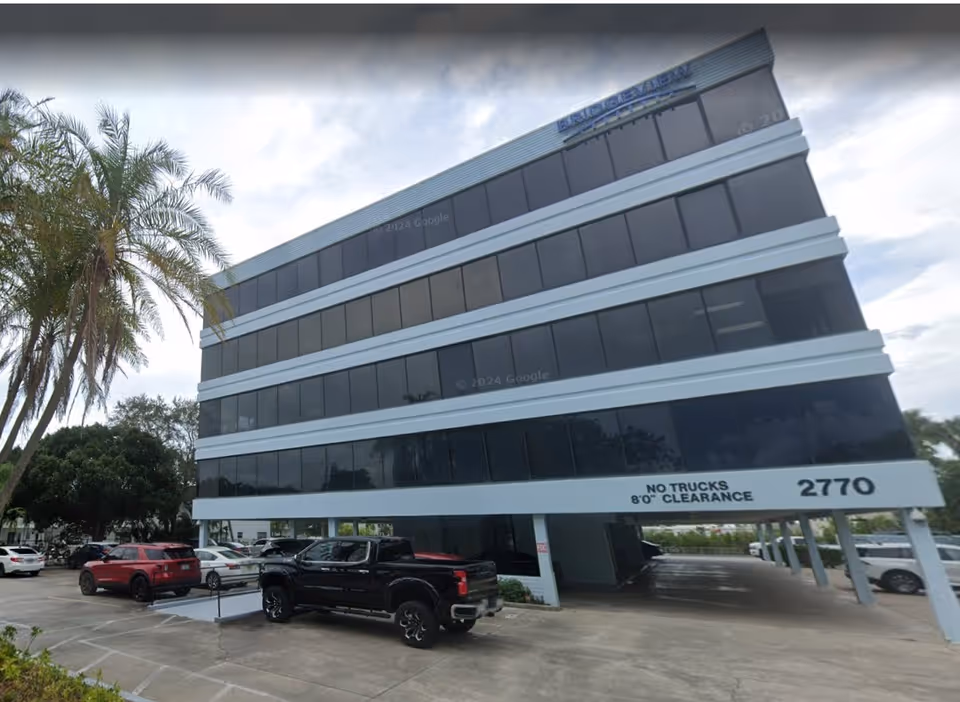Exterior view of a glass, multi-story building with palm trees and parked cars.