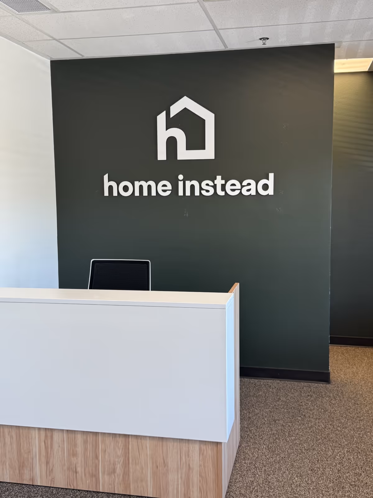 Reception desk in a facility lobby with a dark green wall and Home Instead logo.