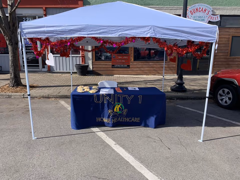 Blue Unity 1 Home Healthcare table sits under white canopy on sidewalk with red garlands.