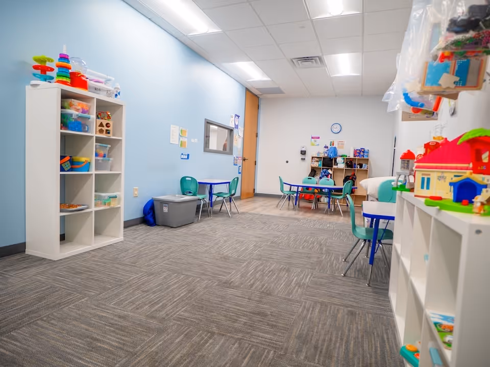 Bright playroom with toy shelves, tables, green chairs, blue walls, and a carpeted floor.