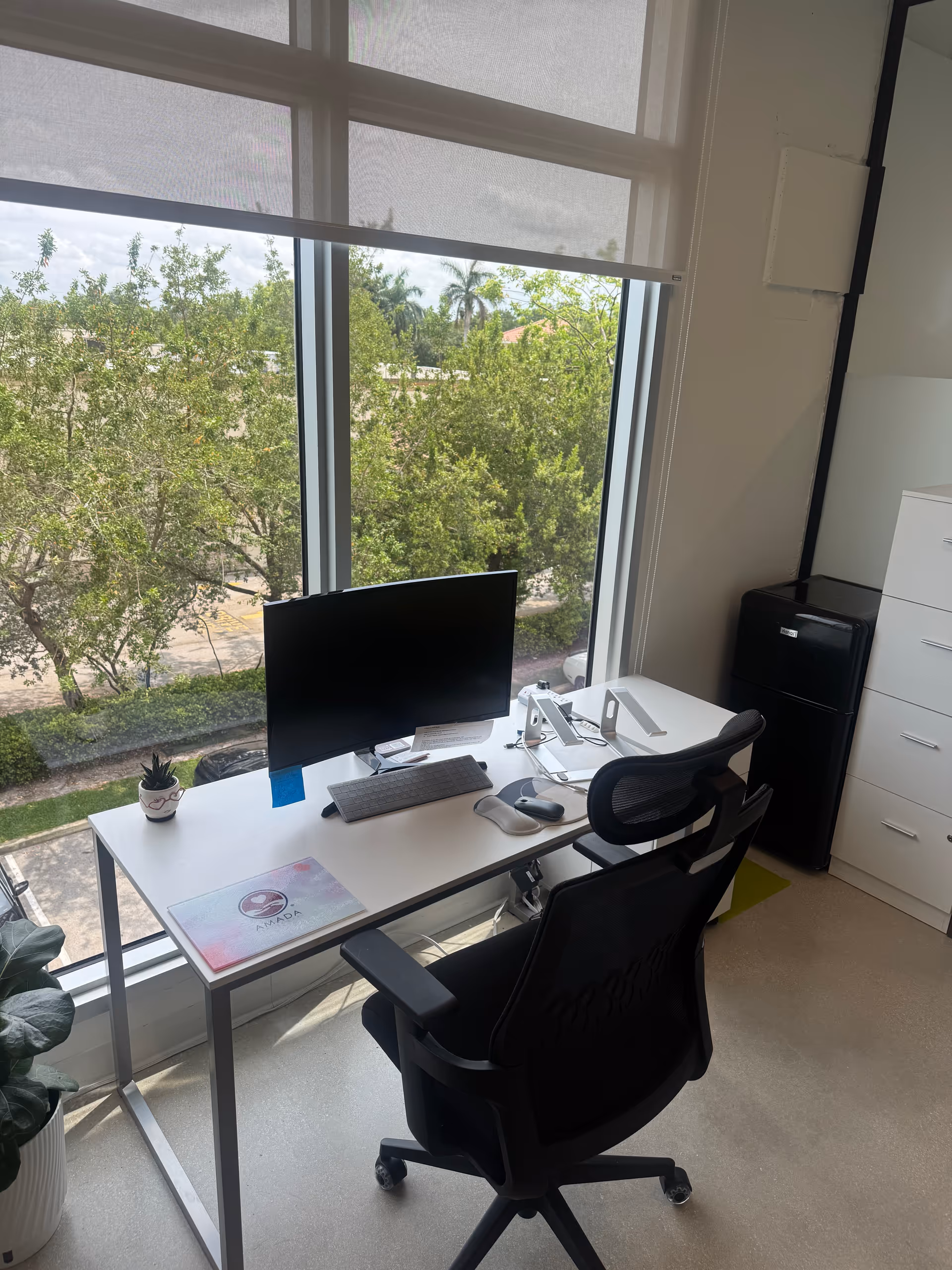 A minimalist office desk by a large window with computer, chair, and storage cabinet.