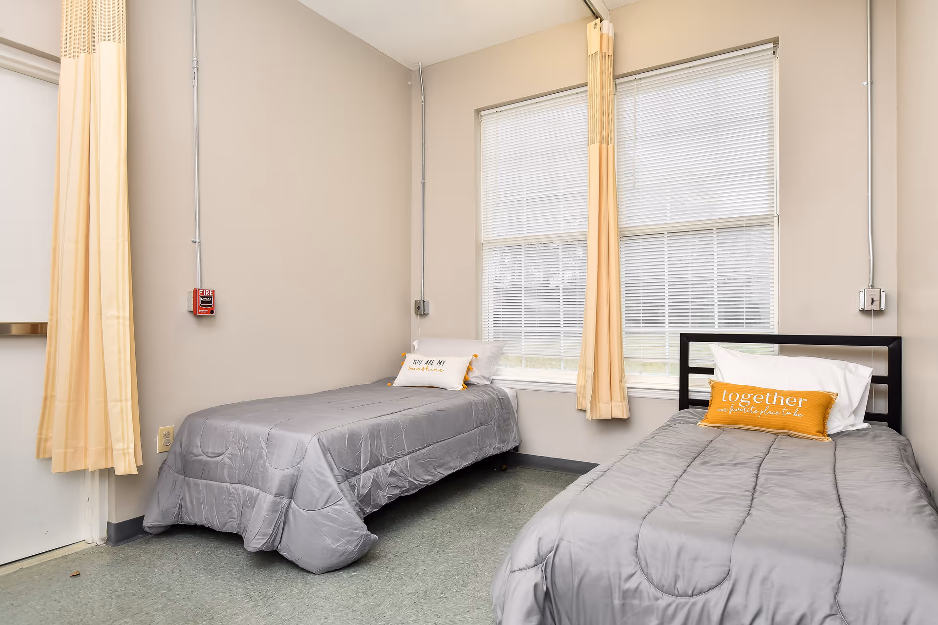 Two single beds with gray quilts sit in a bright shared bedroom with windows and curtains.