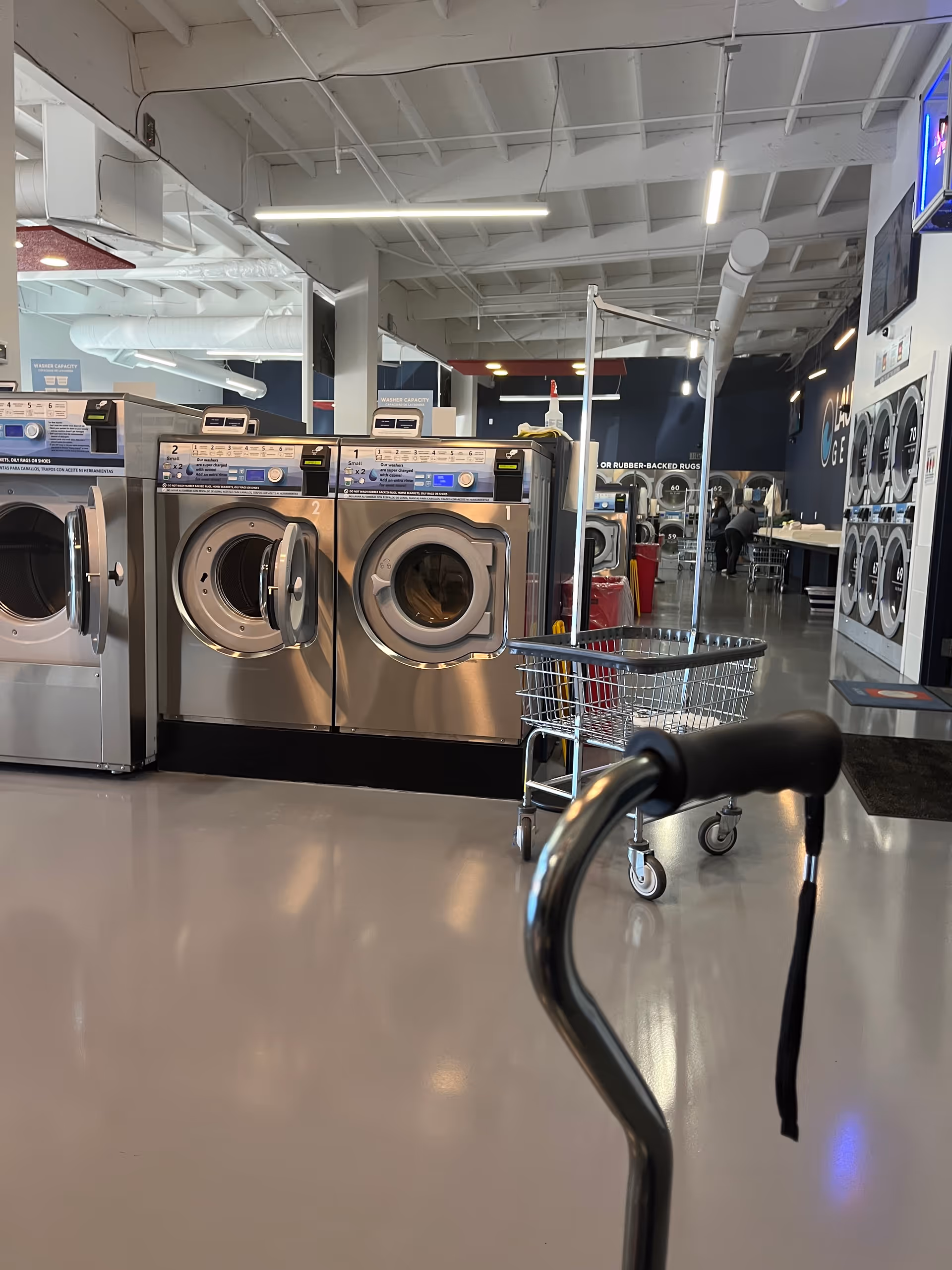 Laundry area with industrial washing machines, a shopping cart, and a walker in a bright facility.