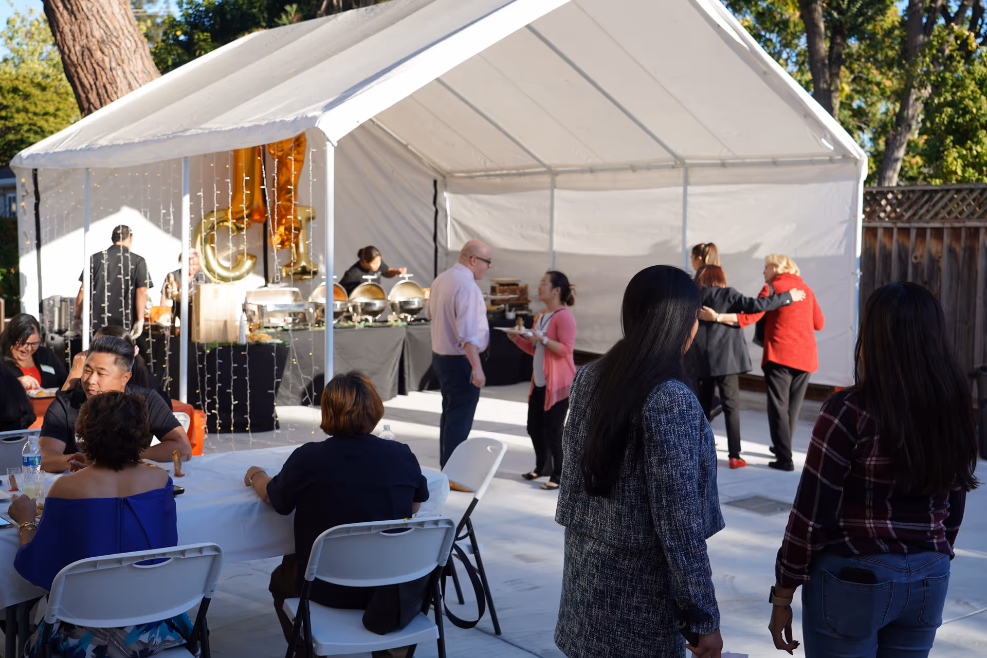 Outdoor event under a white tent with a buffet, seating, and residents mingling.