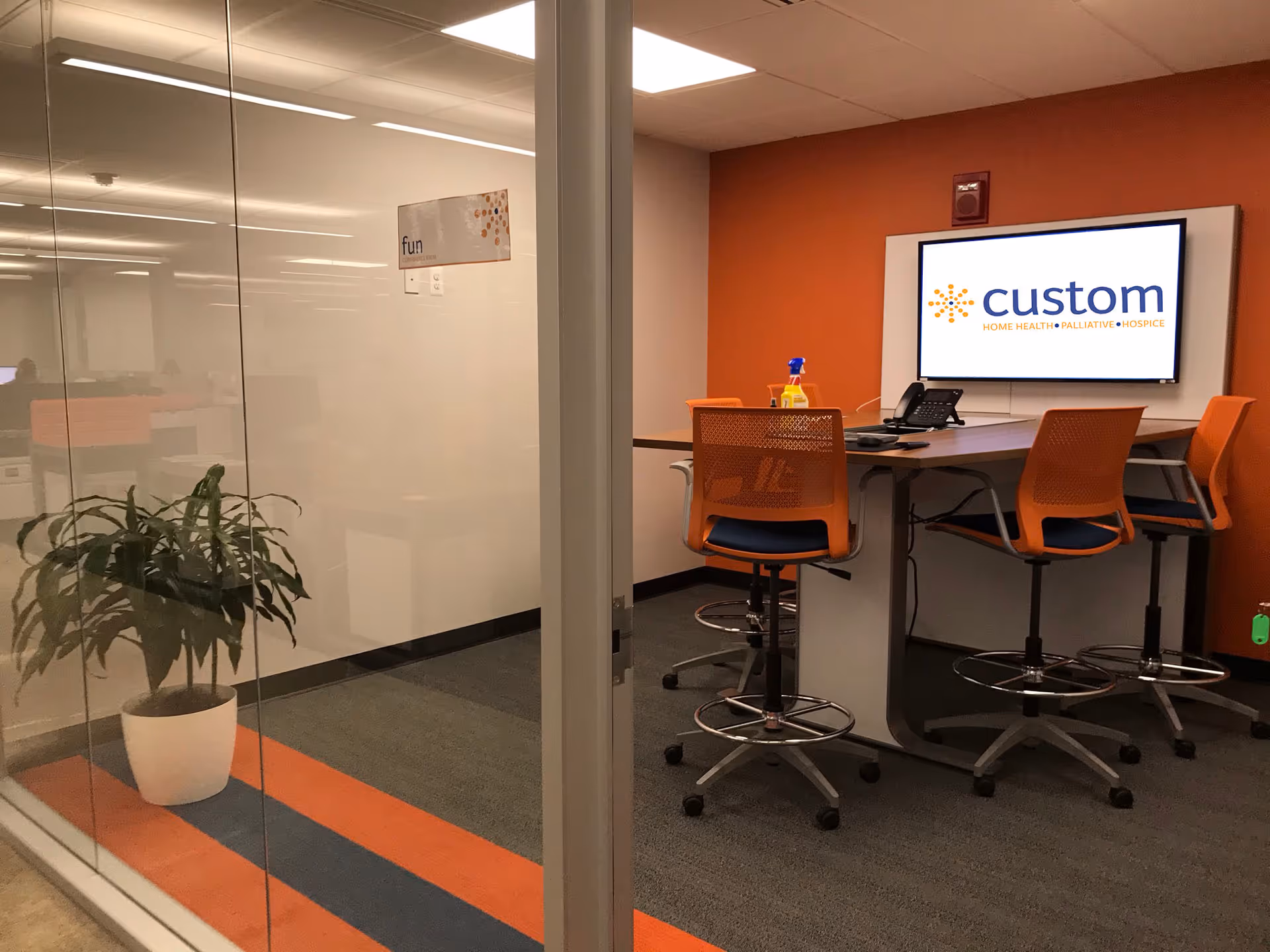 Glass-walled office area with orange chairs around a high table and a Custom logo screen.