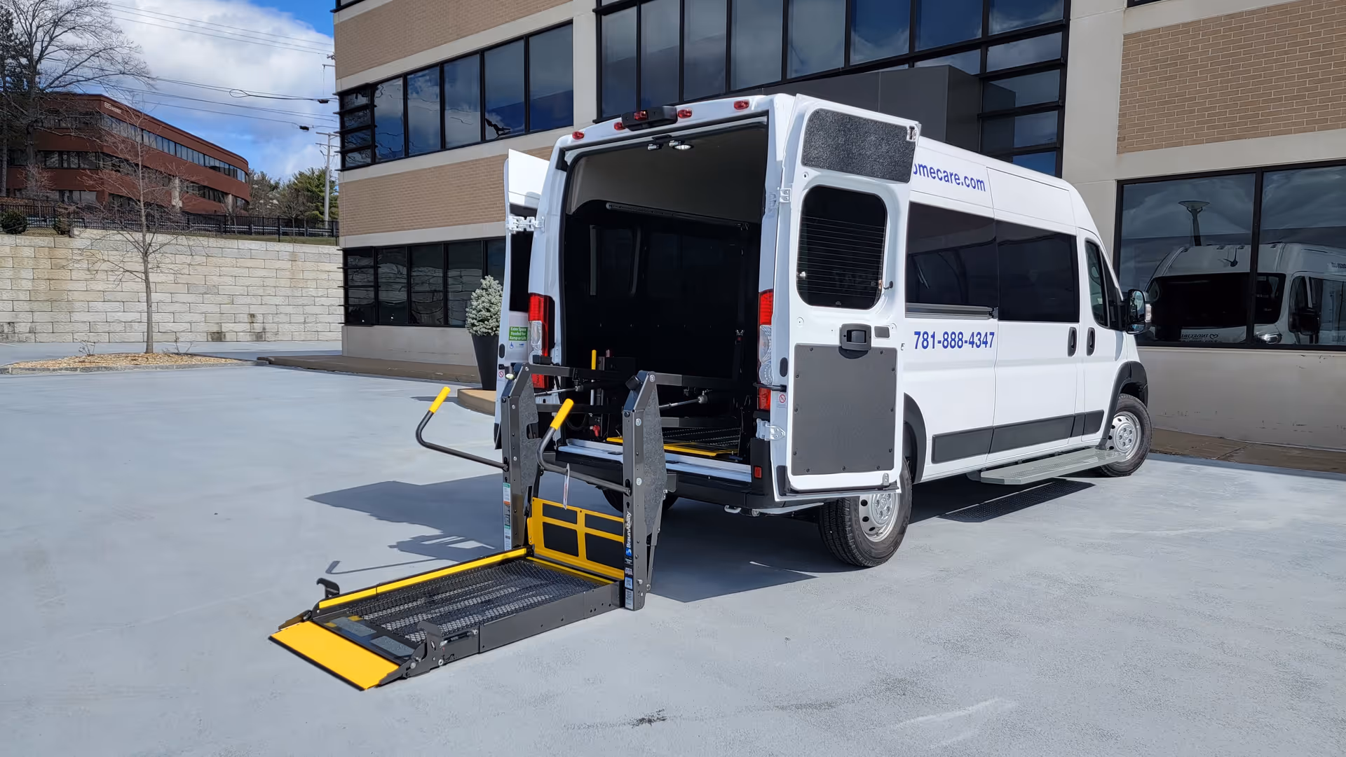 White wheelchair-accessible van with opened ramp parked outside a modern brick and glass building.
