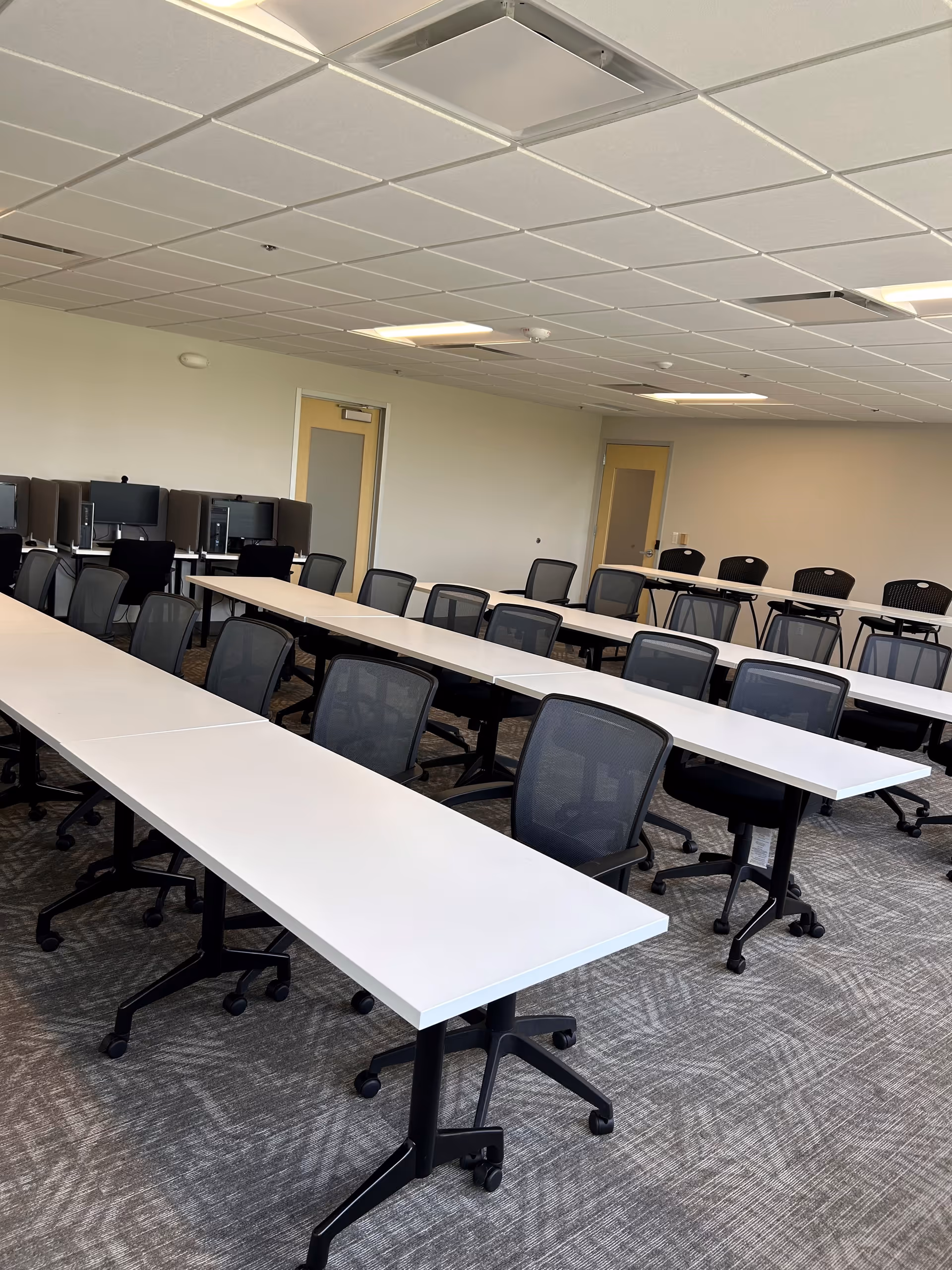 Empty classroom-like room with white tables and rolling black chairs.