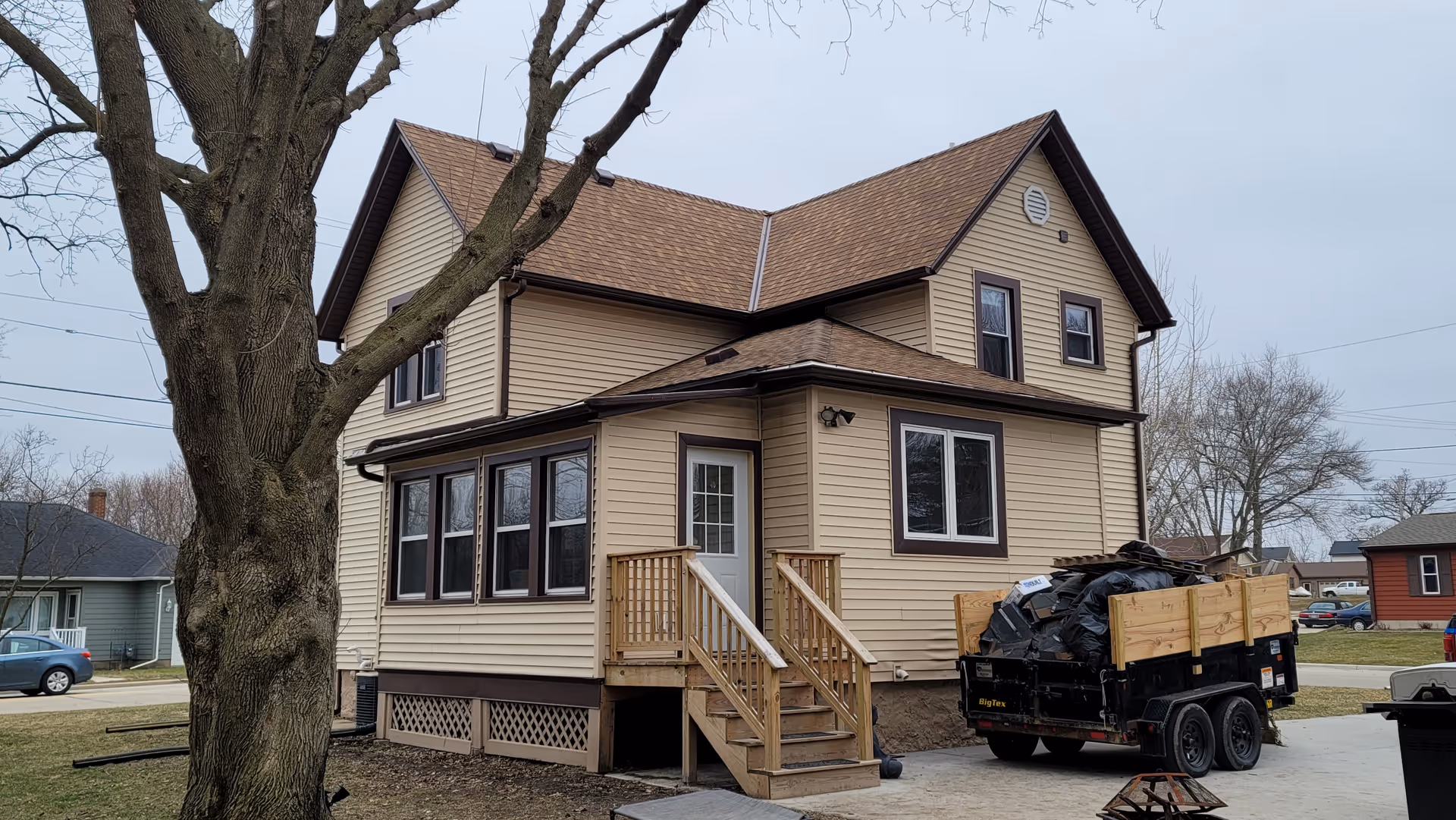 Two-story beige house with wooden stairs and a trailer full of trash bags in driveway