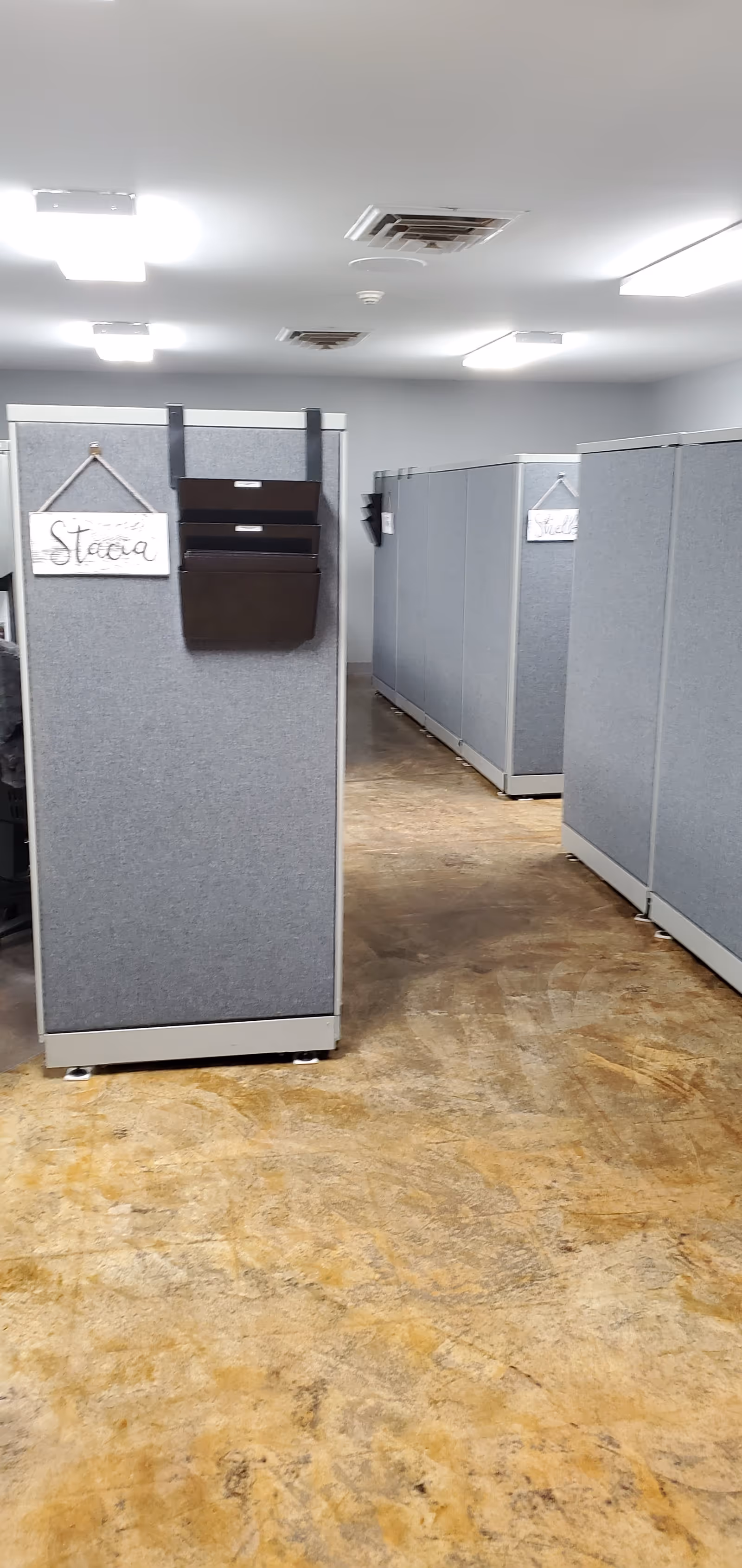 Rows of gray office-style cubicles in a facility hallway with name tags.