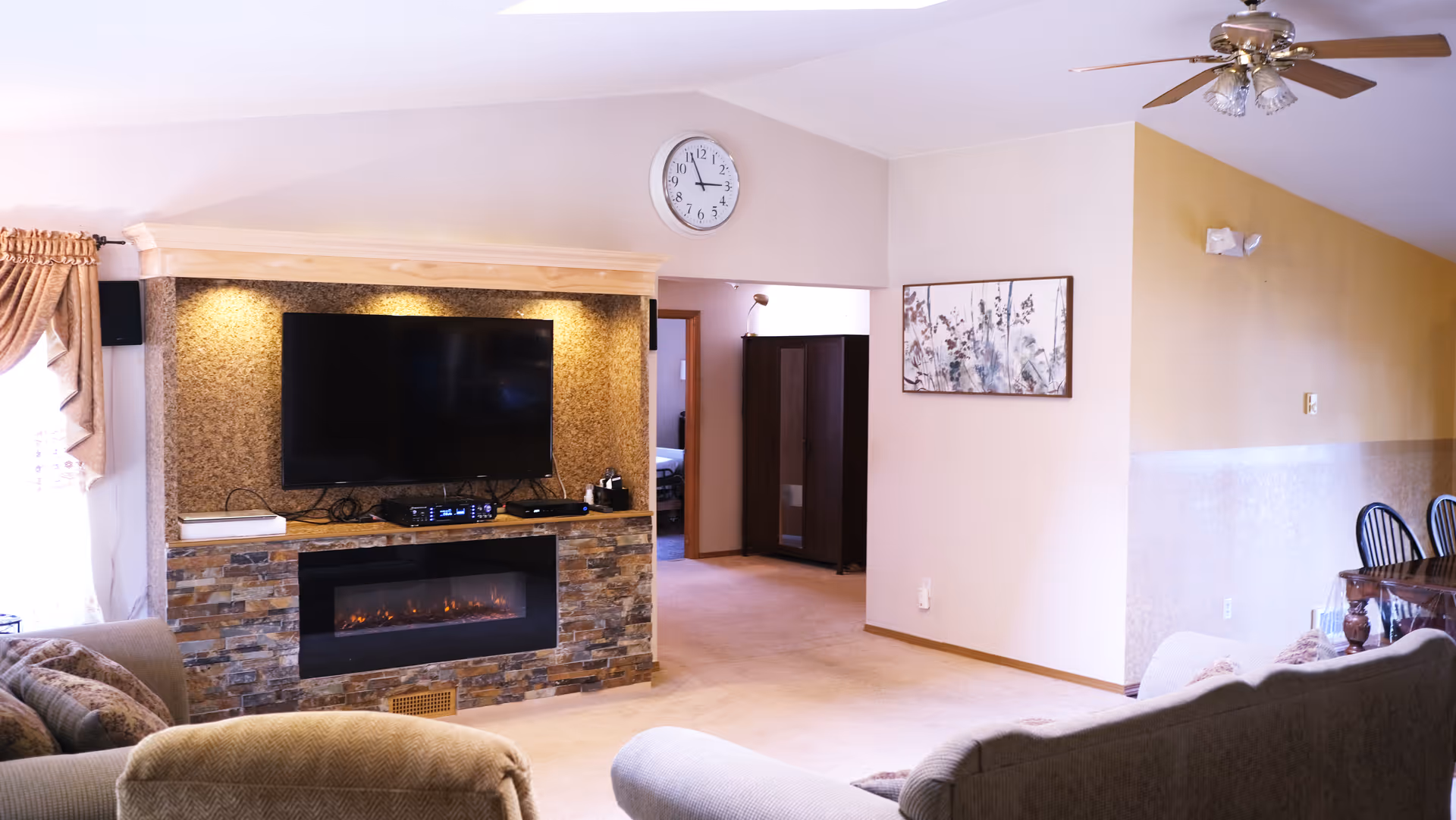 Cozy living room with fireplace, wall TV, clock, sofa, and dining area in a senior home.