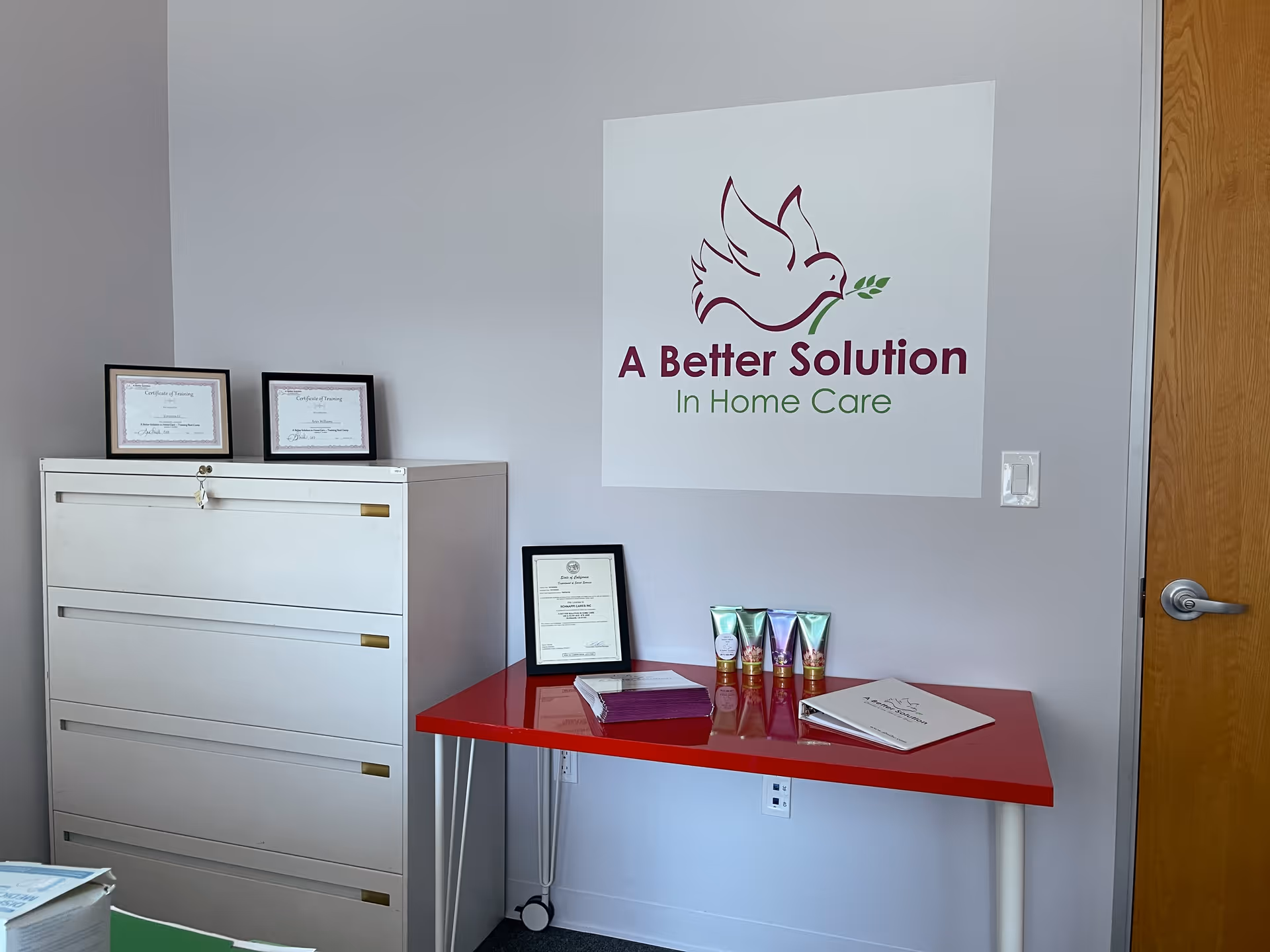 Interior reception area with logo sign, white filing cabinet, red table, and framed certificates.
