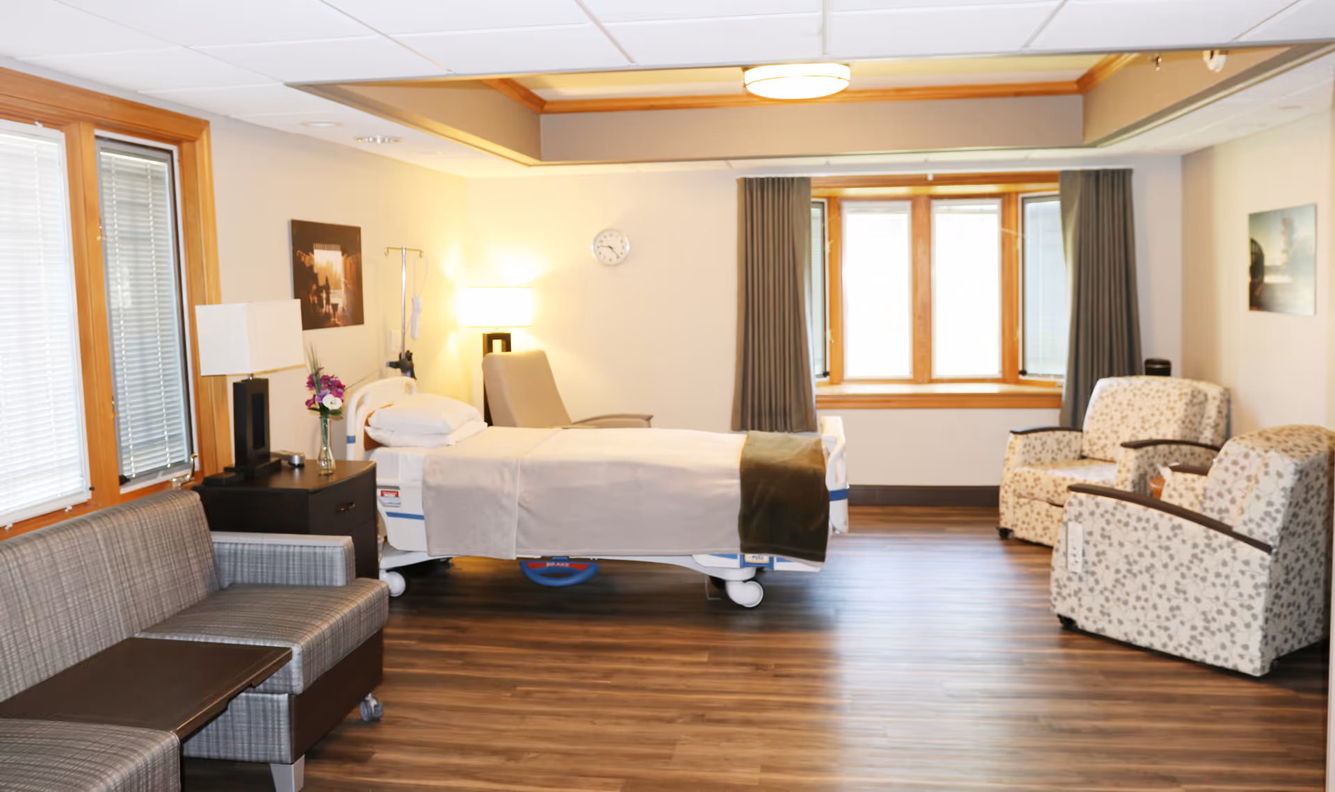 Hospital patient room with a bed, seating, bedside tables, and a windowed view.
