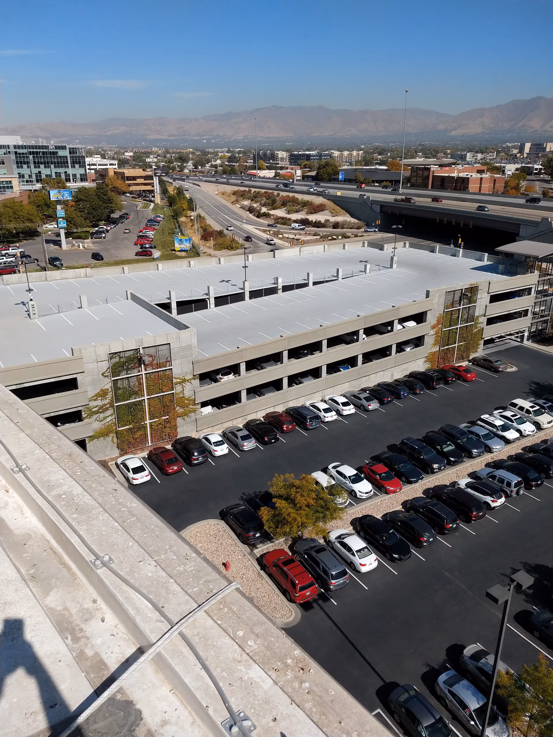Aerial view of a multi-level parking structure beside a highway, with distant mountains.
