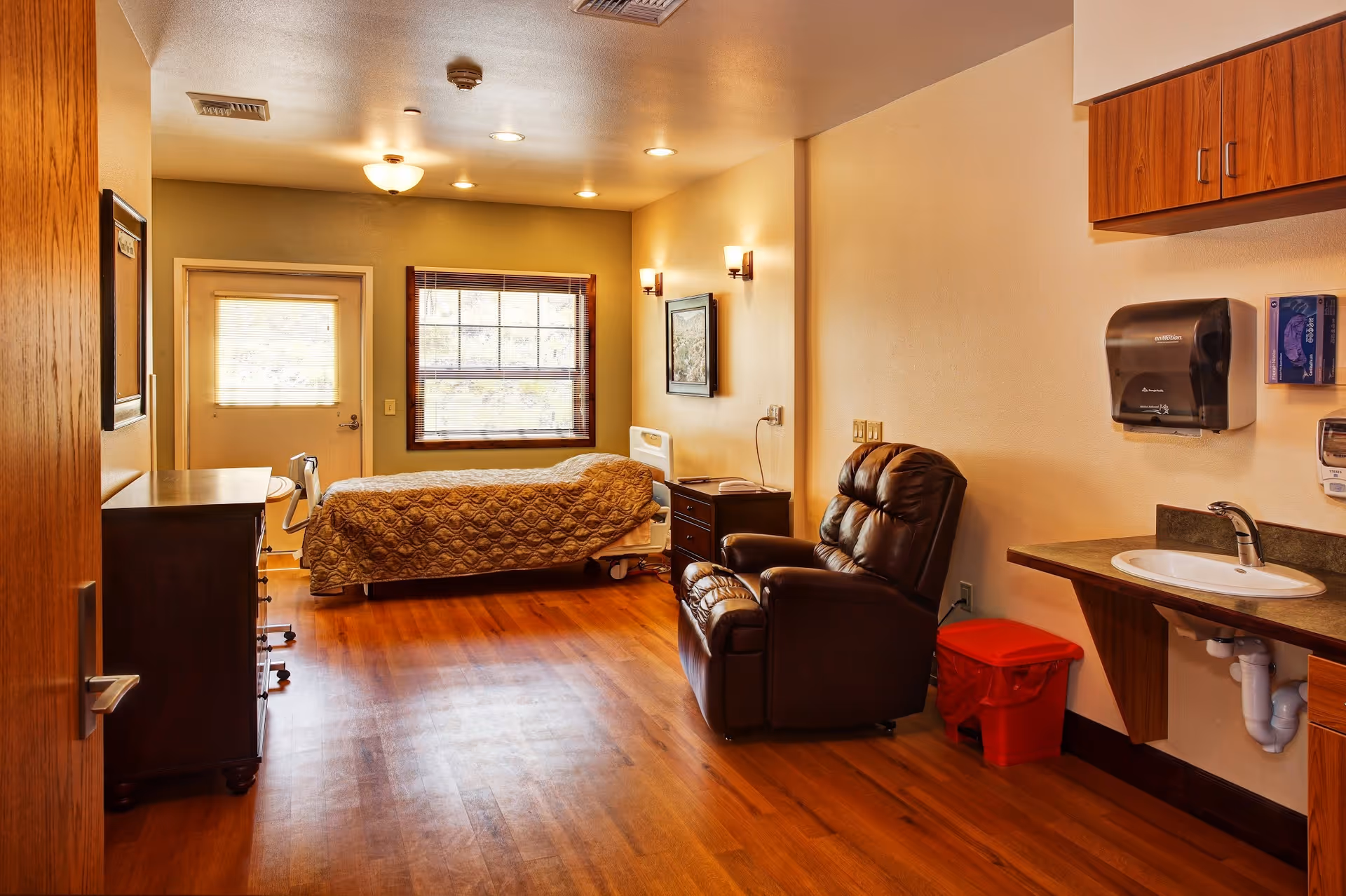Hospice room featuring a bed, brown leather recliner, desk, sink, and wood floors.
