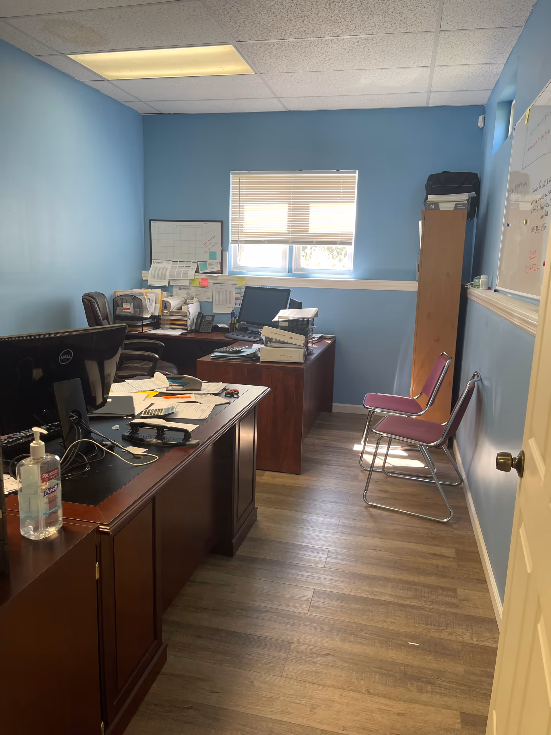 Blue-walled office with desks, computers, papers, and two pink chairs along the wall.