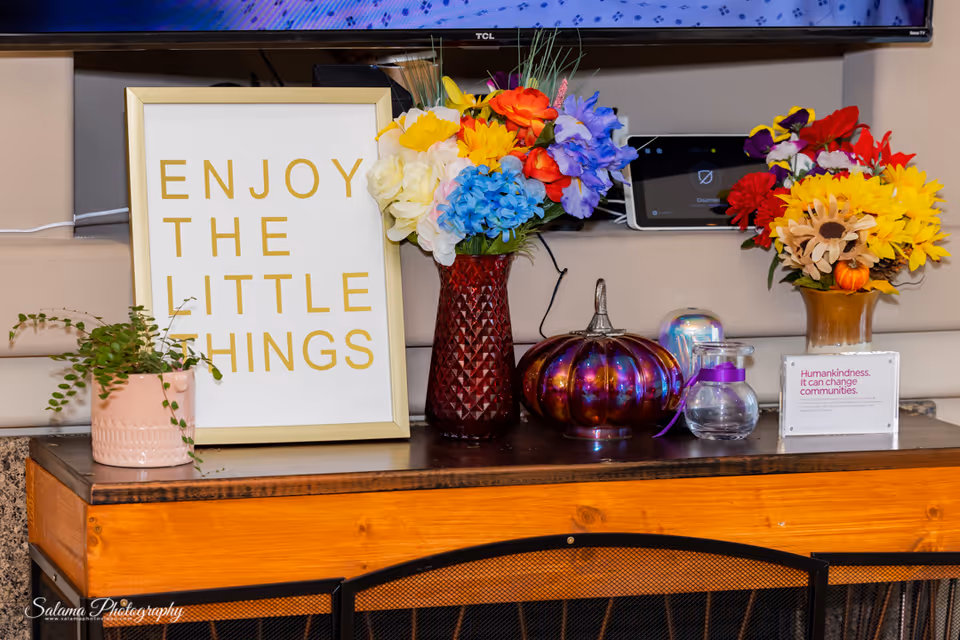 Decorative interior console with colorful bouquets, a framed 'ENJOY THE LITTLE THINGS' sign, and pumpkins.