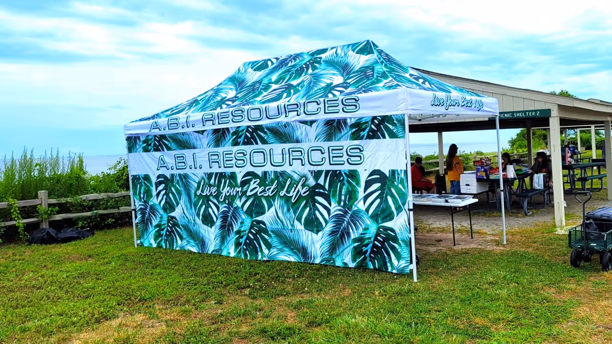 An outdoor scene shows a palm-patterned A.B.I. Resources tent with people near a pavilion.