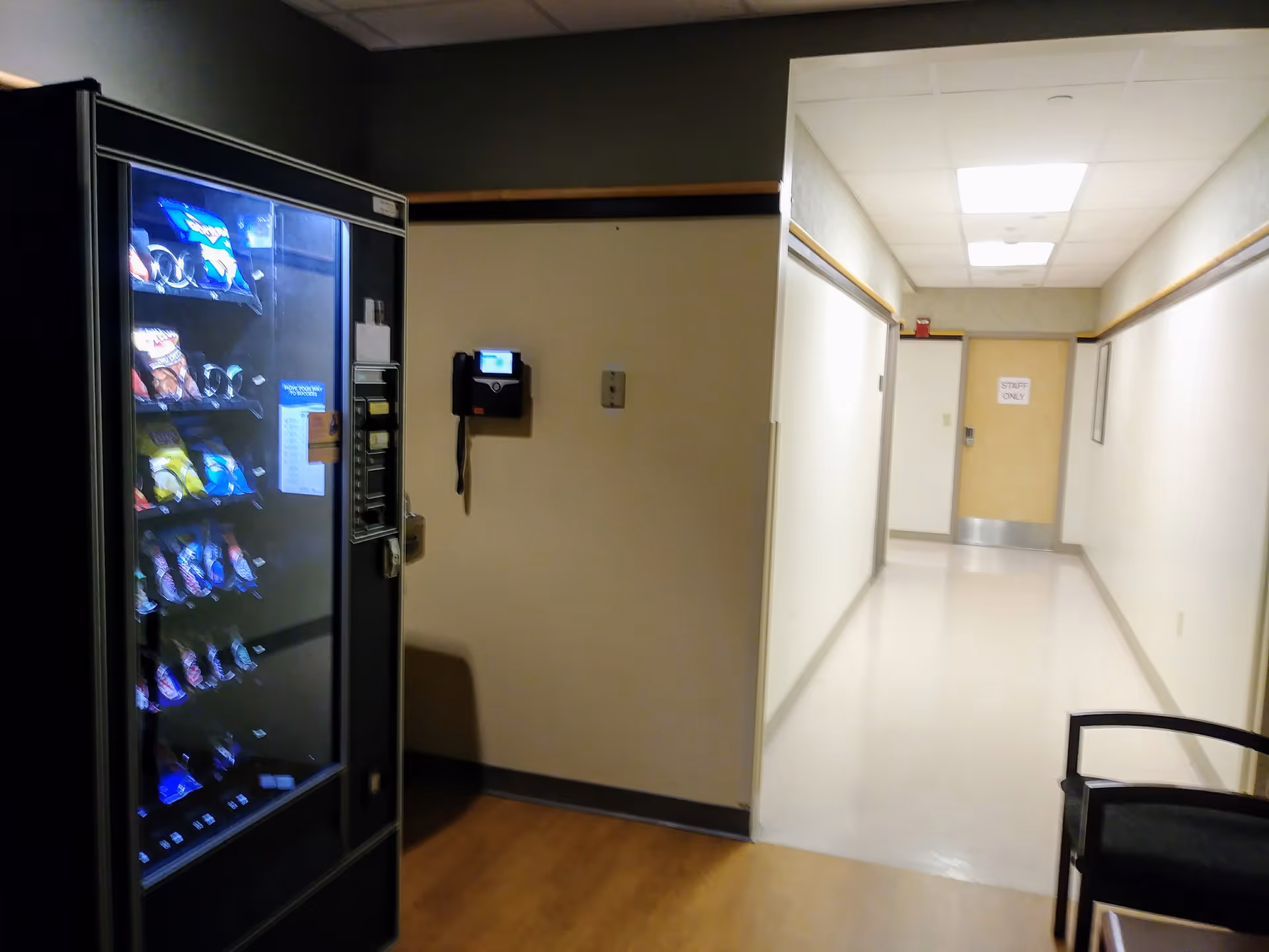 A vending machine sits in a hospital hallway with beige walls and doors.
