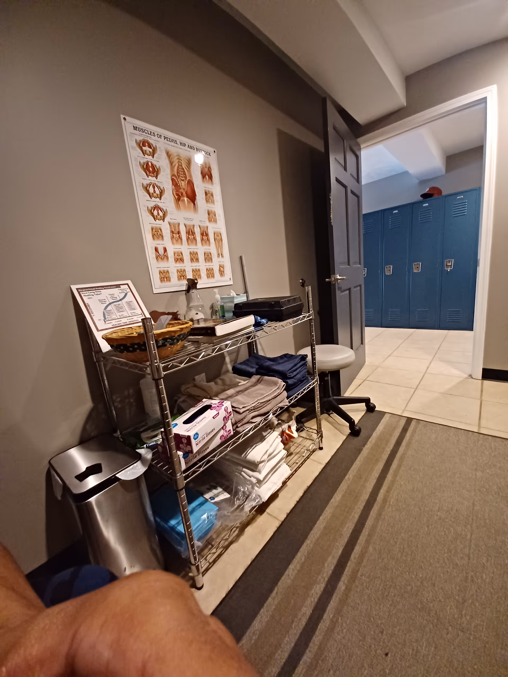 Hallway with metal shelving, medical supplies, and blue lockers at an open door.
