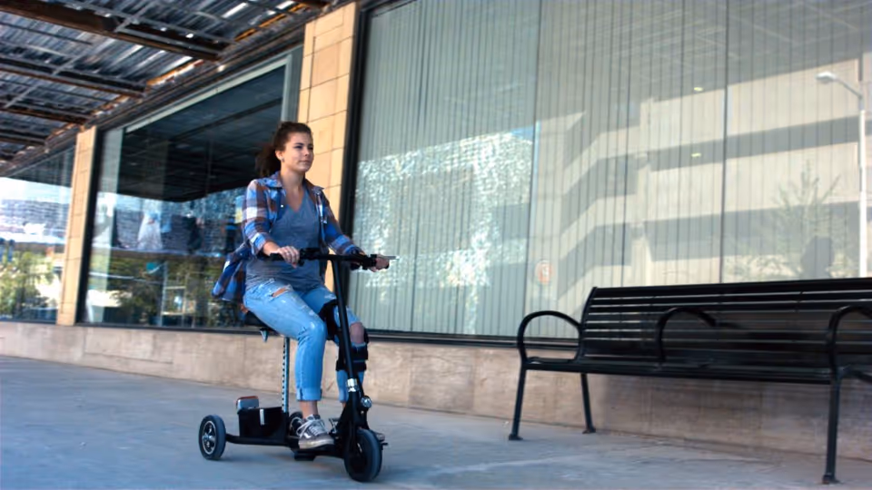 Young woman rides a small electric scooter outside a glass-fronted building with a bench.