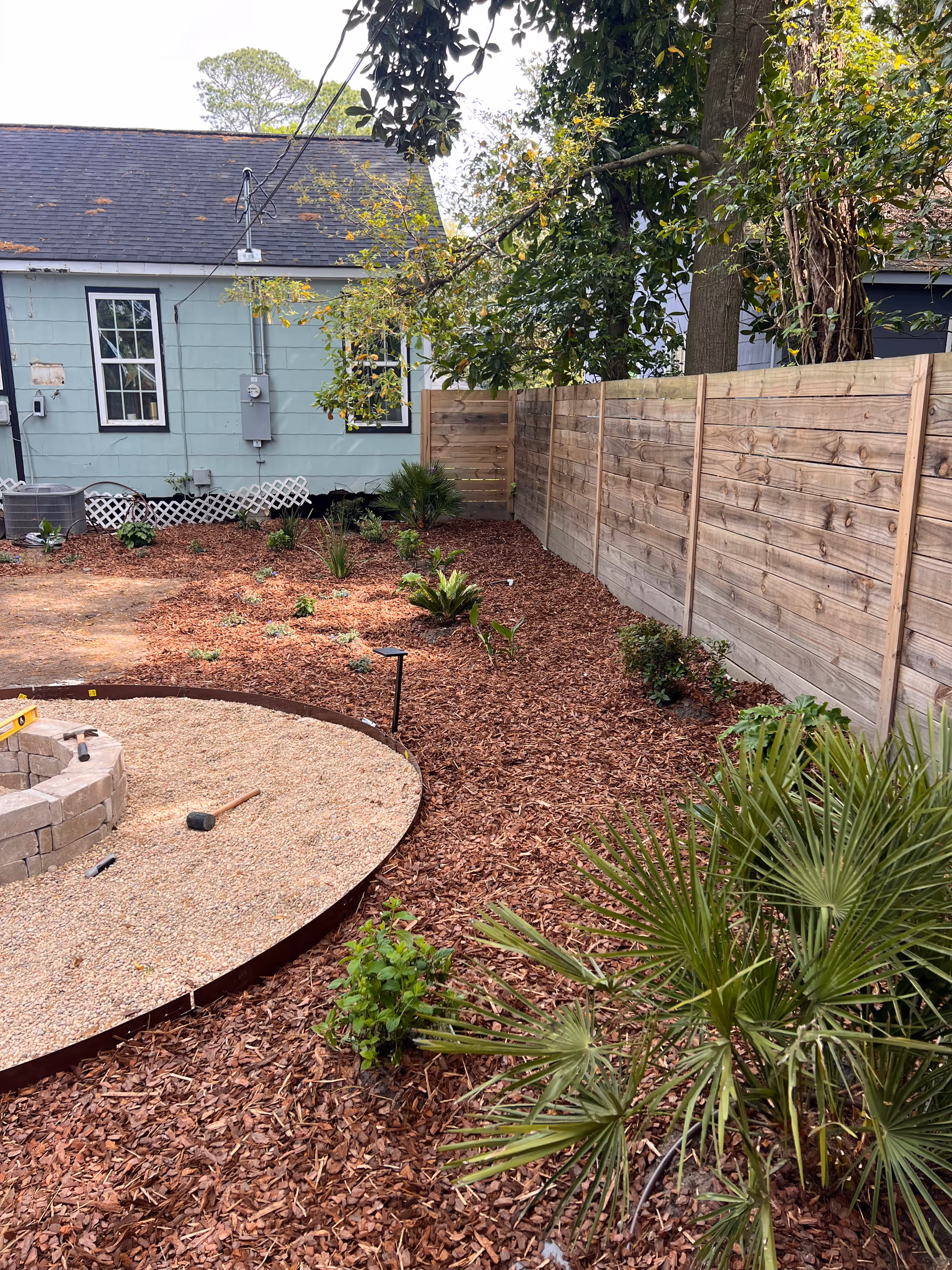 Backyard garden with mulch, a circular fire pit ring, and a wooden privacy fence.