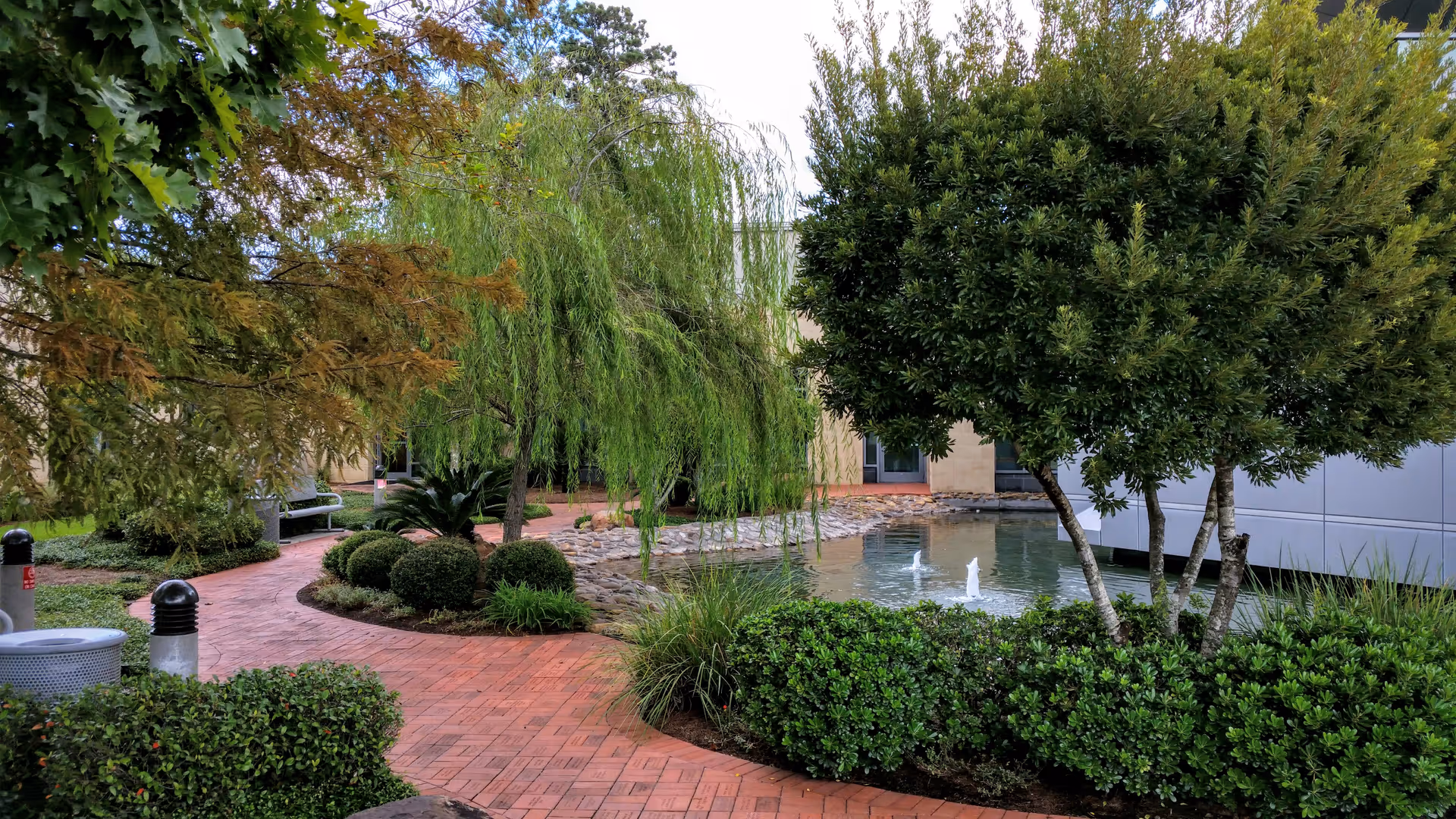Exterior courtyard at the facility with brick walkway, manicured shrubs, trees, and a water fountain.