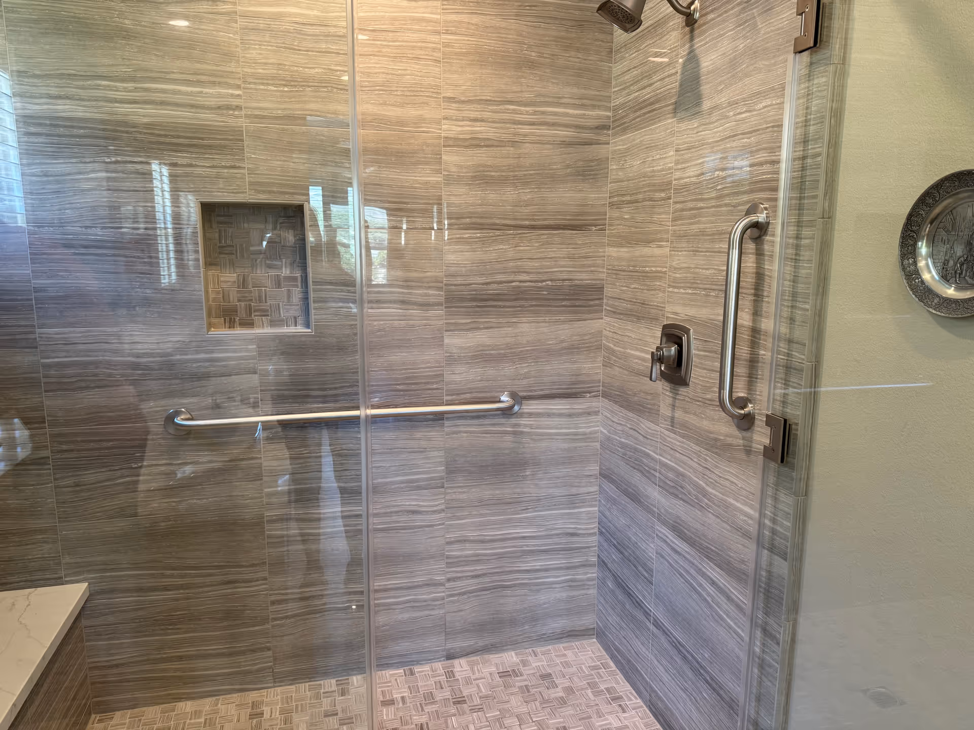 Accessible glass-enclosed shower with gray tile walls, grab bars, and a built-in niche.