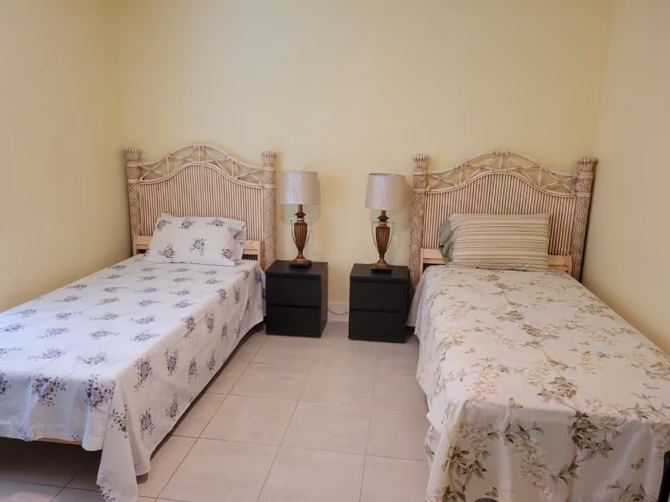 Two single beds with wicker headboards, matching nightstands, and lamps between in a sunny bedroom.