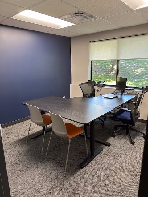 A conference-style office with a long dark table, chairs, computers, and a window.