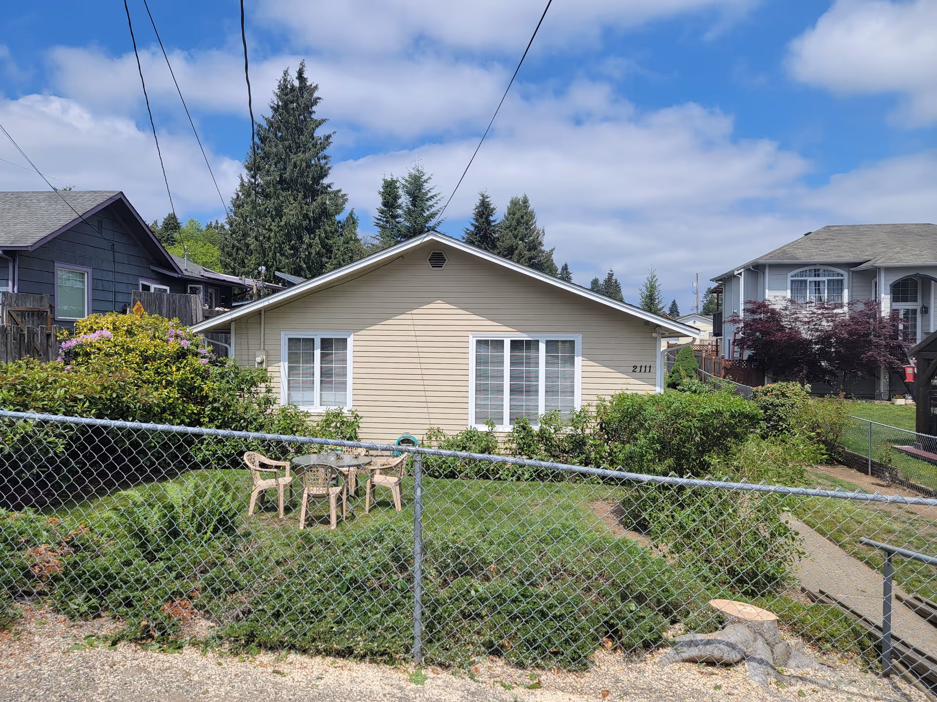 Single-story beige house with a chain-link fenced garden and plastic outdoor table and chairs.