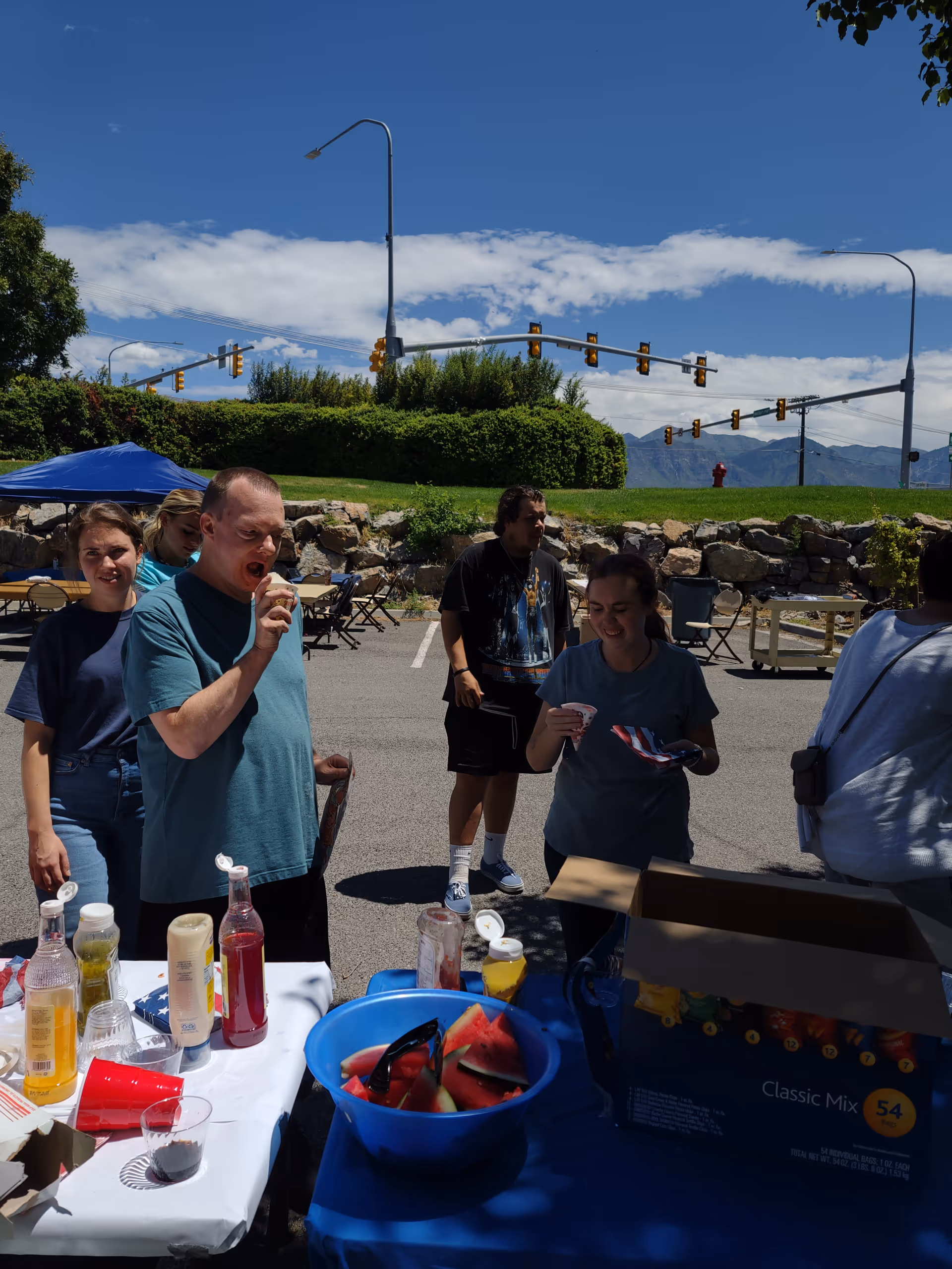 People gather around a table with condiments and watermelon outdoors under a blue sky.