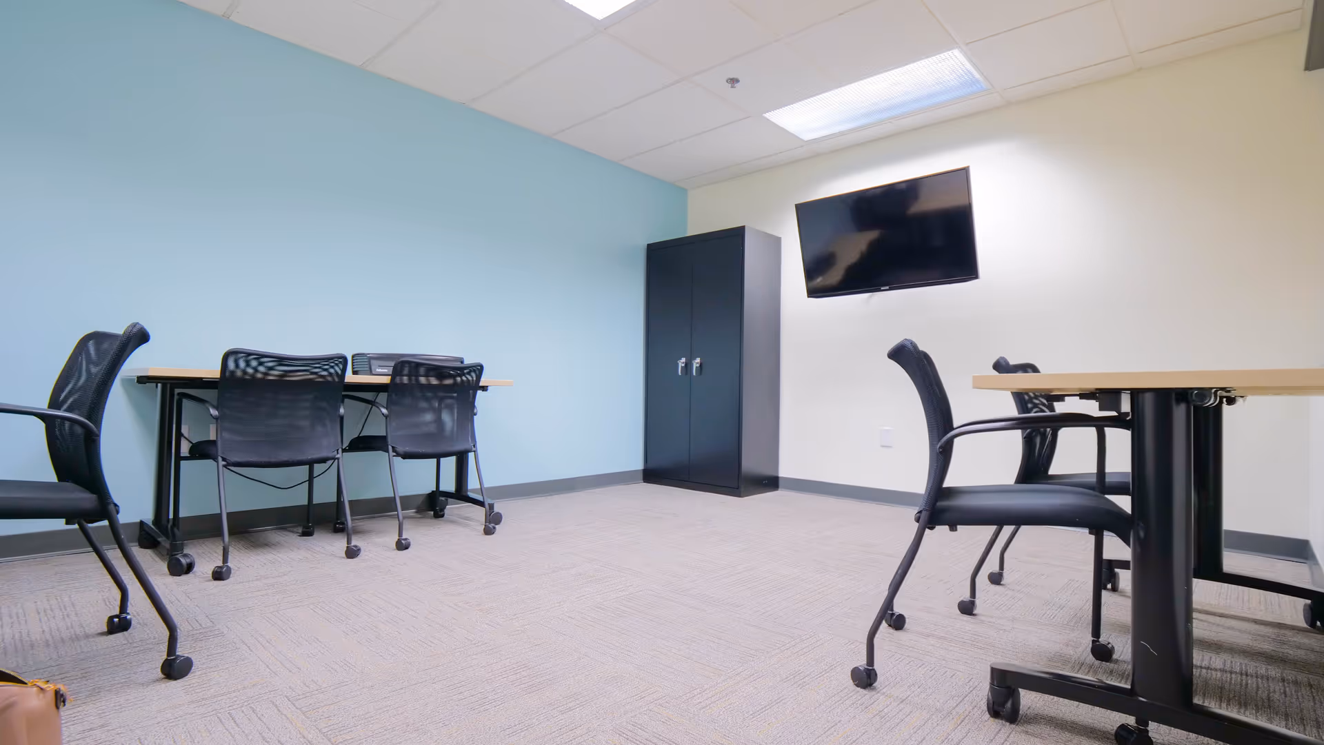 A bright, empty conference room with tables, chairs, a cabinet, and a wall-mounted TV.