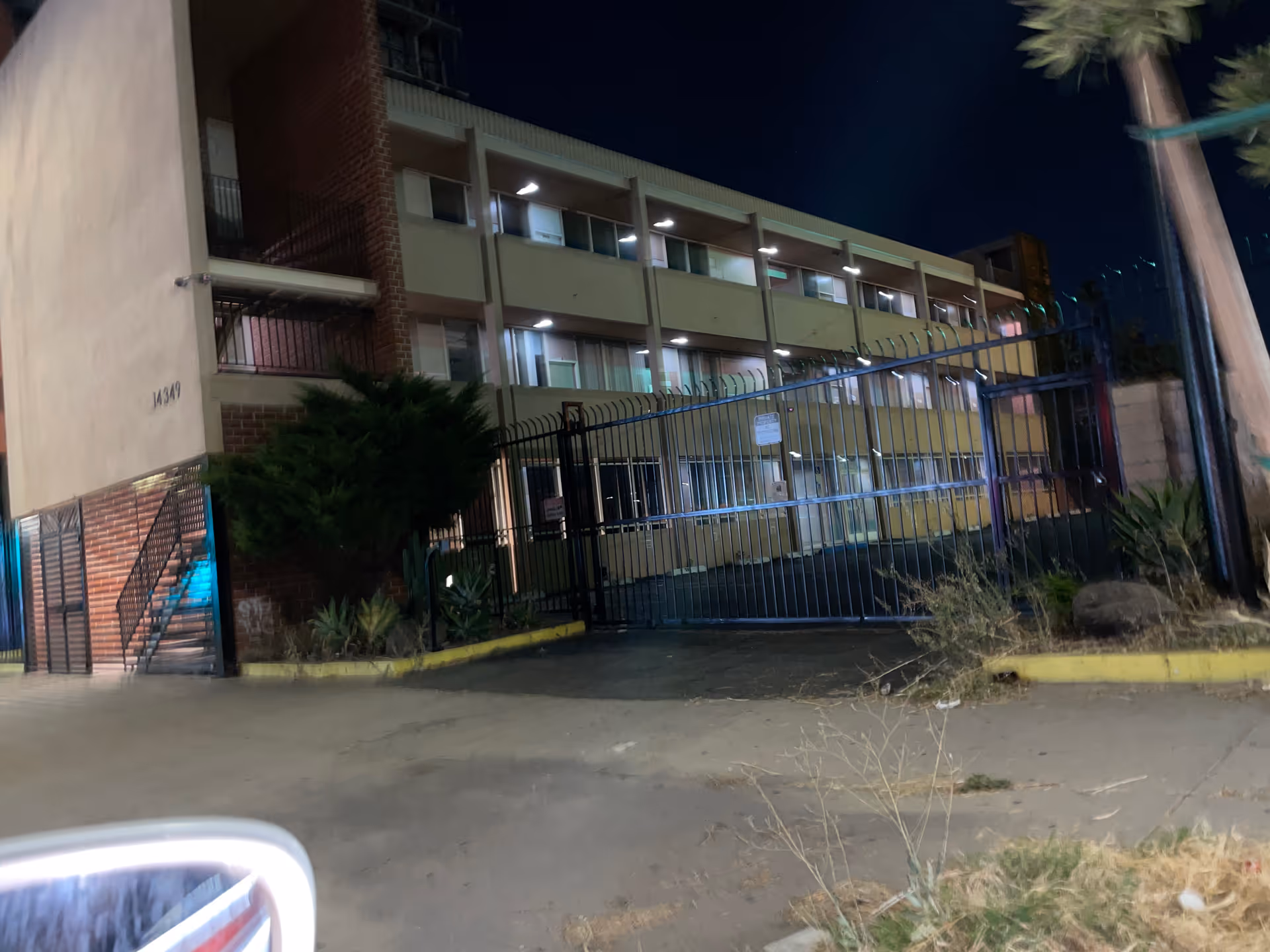Night exterior view of a multi-story gated facility with a tall metal fence.