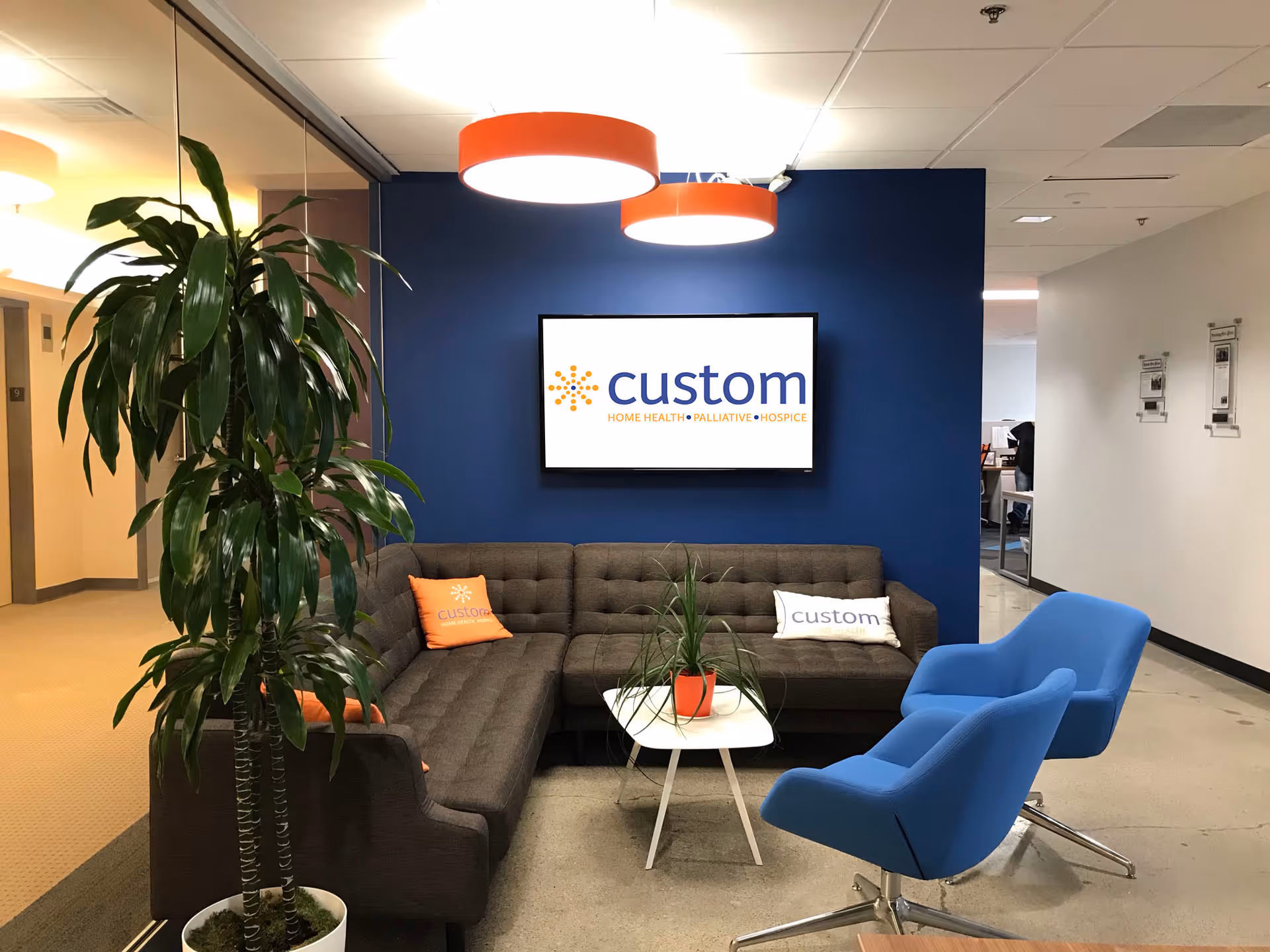 Lobby seating area with blue wall, mounted screen displaying custom, sofa, and bright orange chairs.