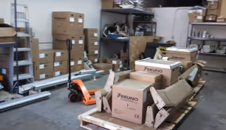 Cluttered storage room with shelves, cardboard boxes labeled BRUNO, and a pallet jack.