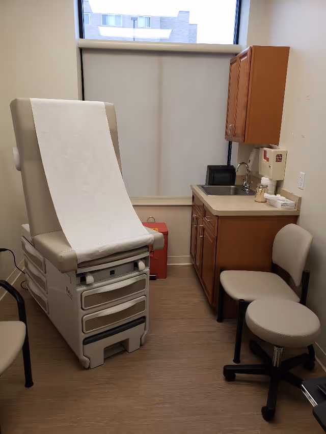 Medical examination room with a paper-covered exam chair, sink, cabinets, and stools.