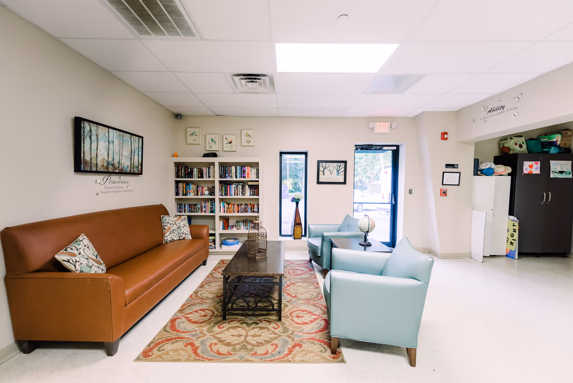 A cozy senior living common area with a brown sofa, teal chairs, and a bookshelf.
