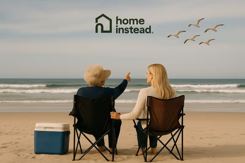 Two people sit in folding beach chairs on the shore beneath Home Instead branding.