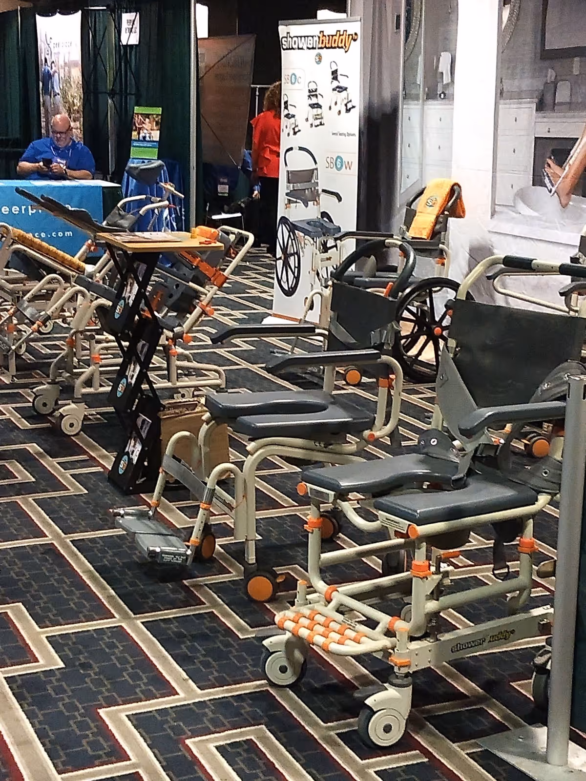 Indoor exhibit area with walkers, wheelchairs, and chairs; a man browses near banners.
