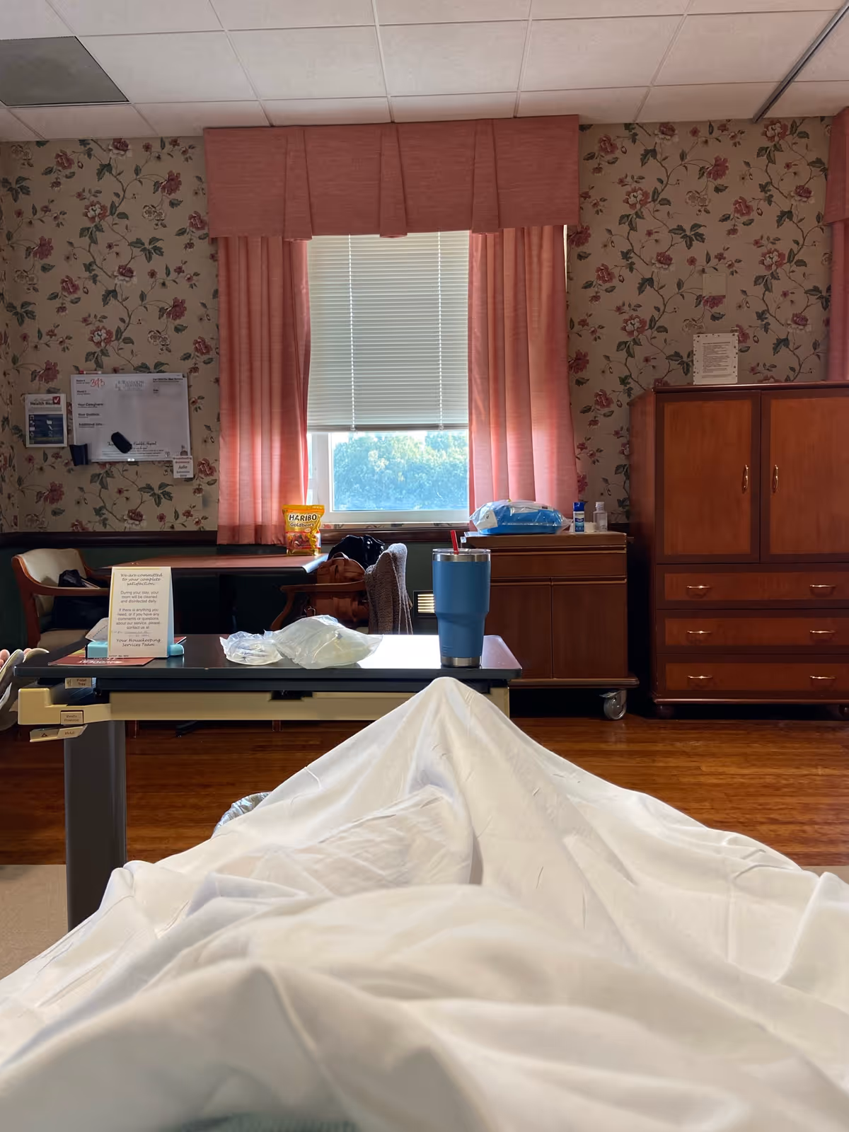 Hospital-style patient room with a bed covered by a white sheet, pink curtains.