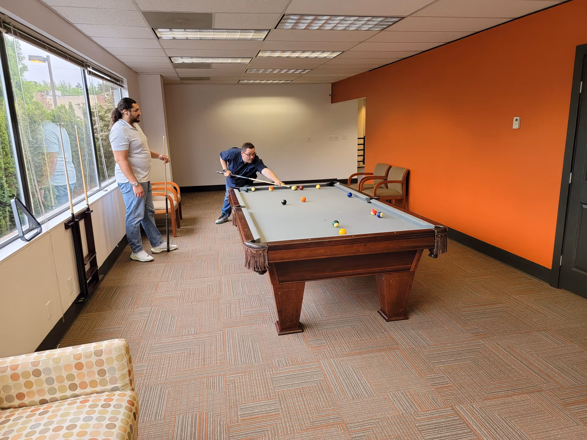 Two men play billiards in a recreation room with orange accent wall.