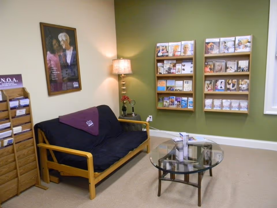 A small waiting lobby with a wooden sofa, glass coffee table, lamp, and magazine racks.