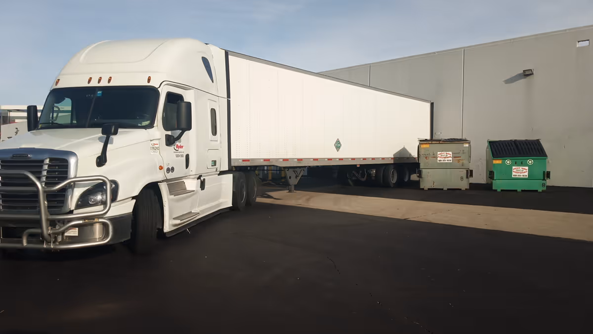 A white semi-truck with a long trailer parked beside a large gray building, dumpsters nearby.