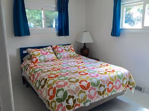 Bright bedroom with colorful heart-pattern bedspread, two windows, and a nightstand lamp.