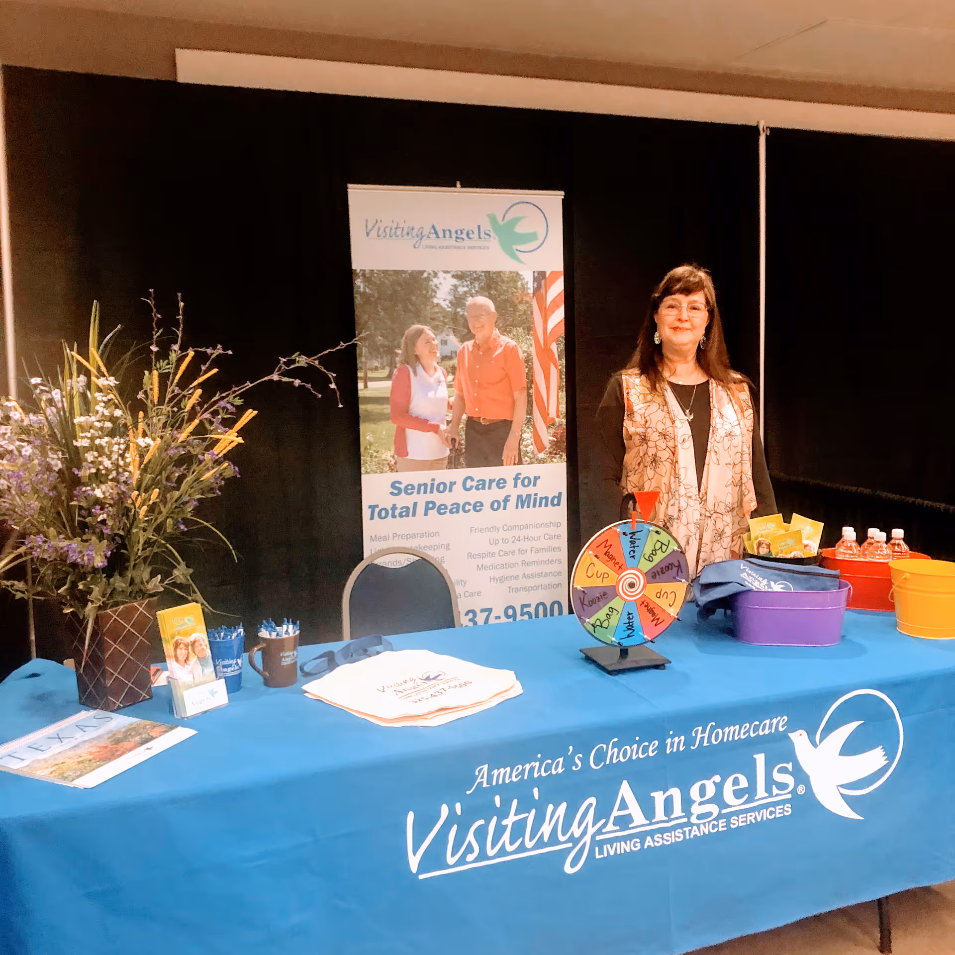 Promotional booth for Visiting Angels Senior Home Care indoors with banner, table, and a standing representative.