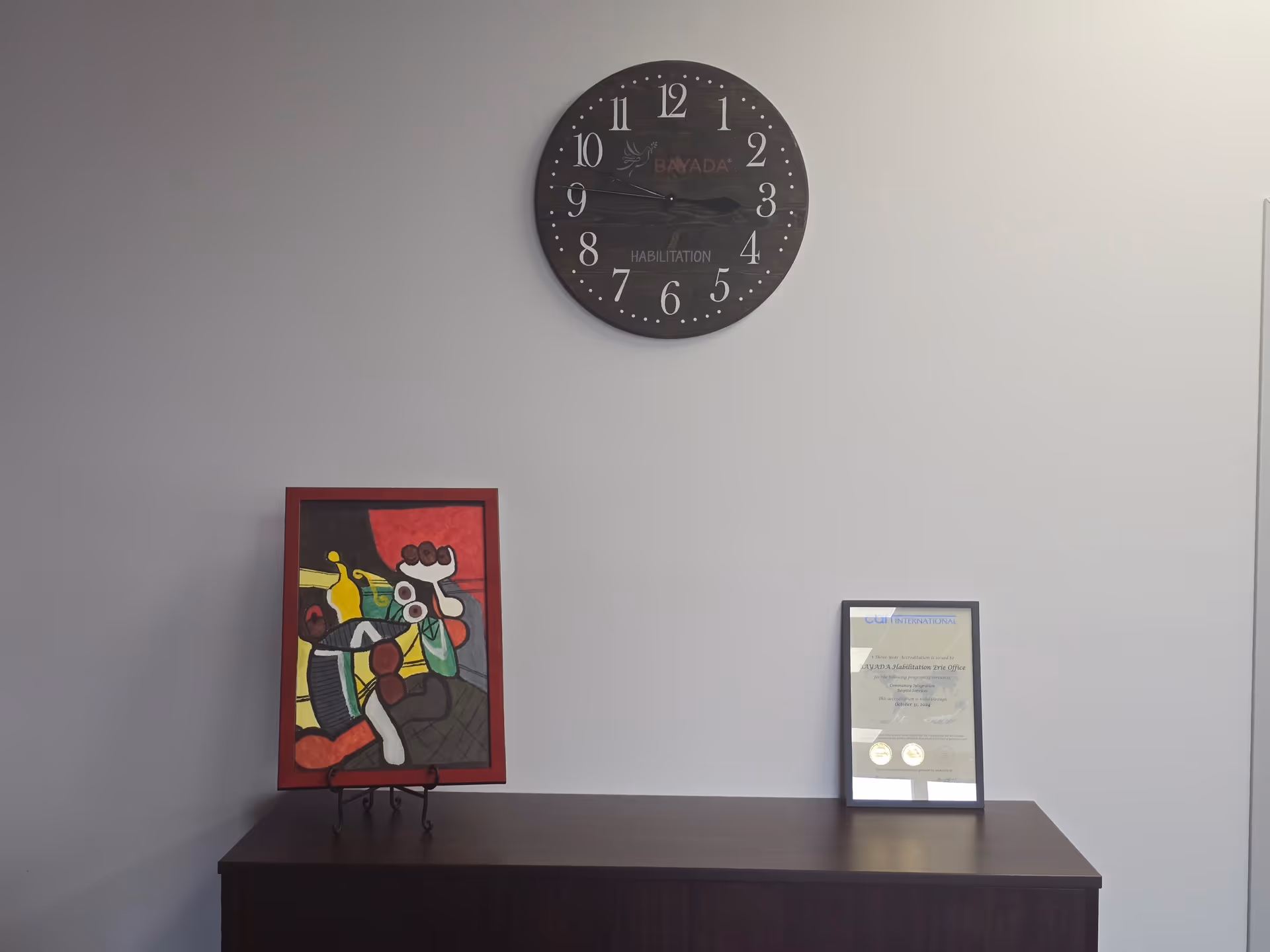 Gray wall with a large clock above a dark cabinet, plus framed art and a certificate