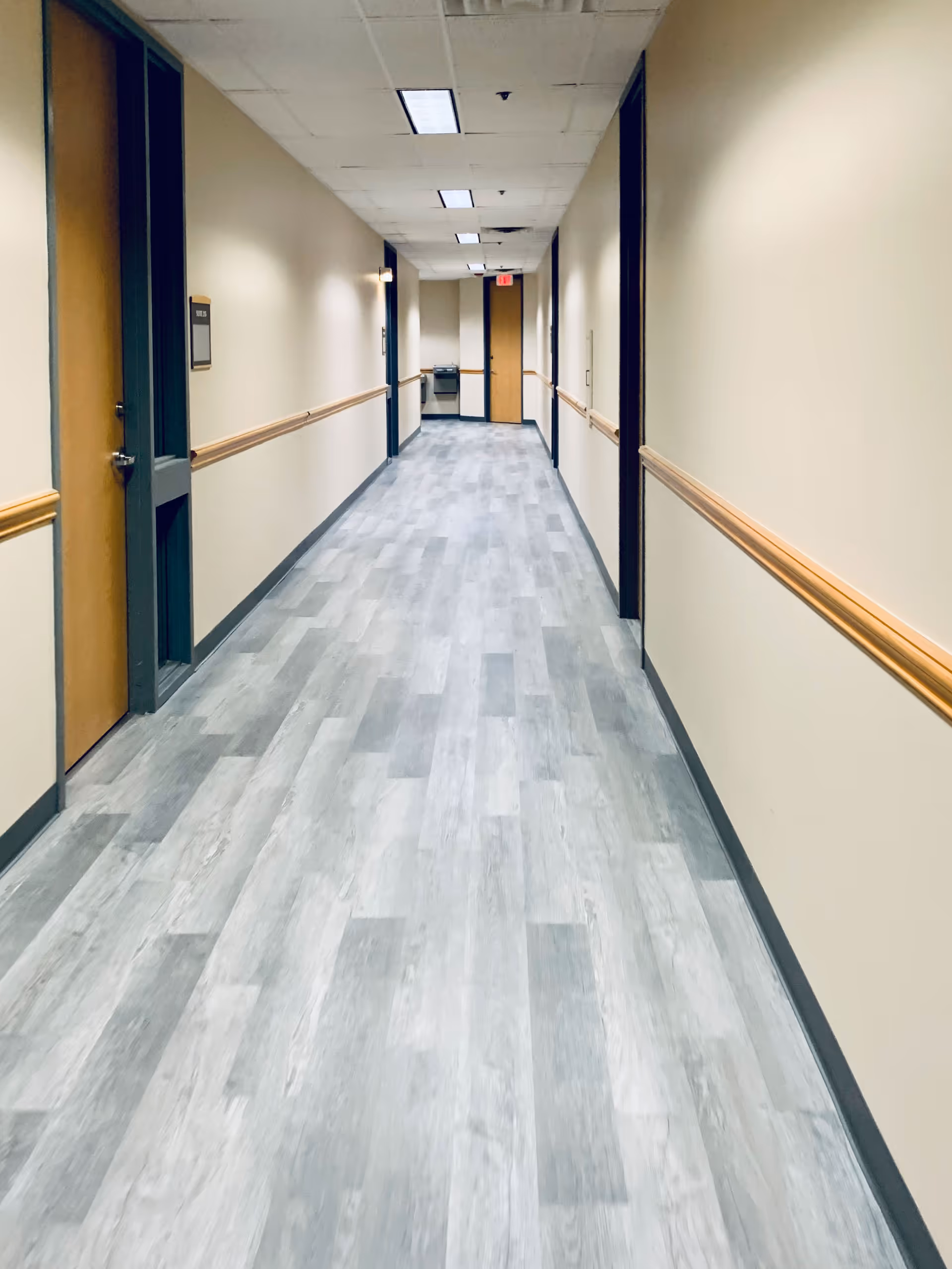 A long, empty hospital hallway with doors, beige walls, and blue-gray flooring.